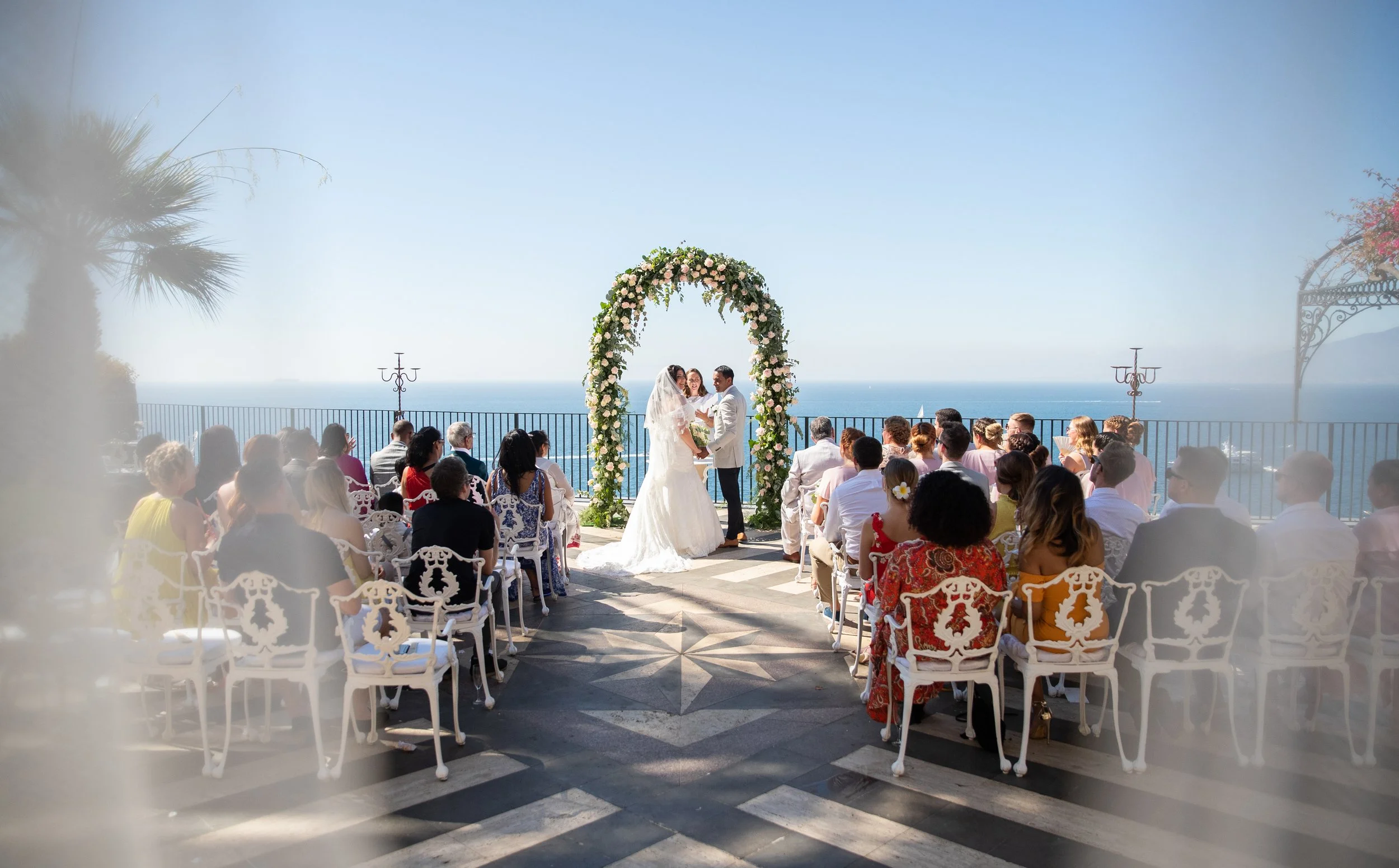 A wedding ceremony taking place outdoors on a terrace overlooking the ocean, with guests seated and a couple exchanging vows under a floral arch.