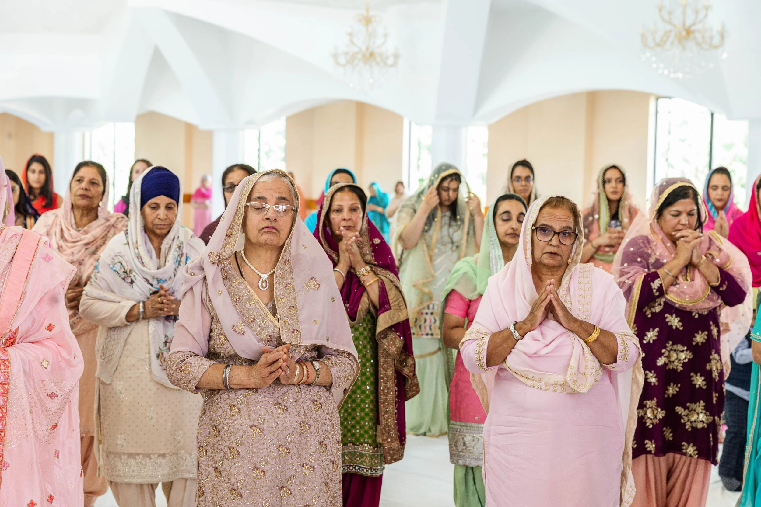 A group of women dressed in traditional colorful Indian attire praying with hands folded in a ceremony inside a bright hall with chandeliers and large windows.