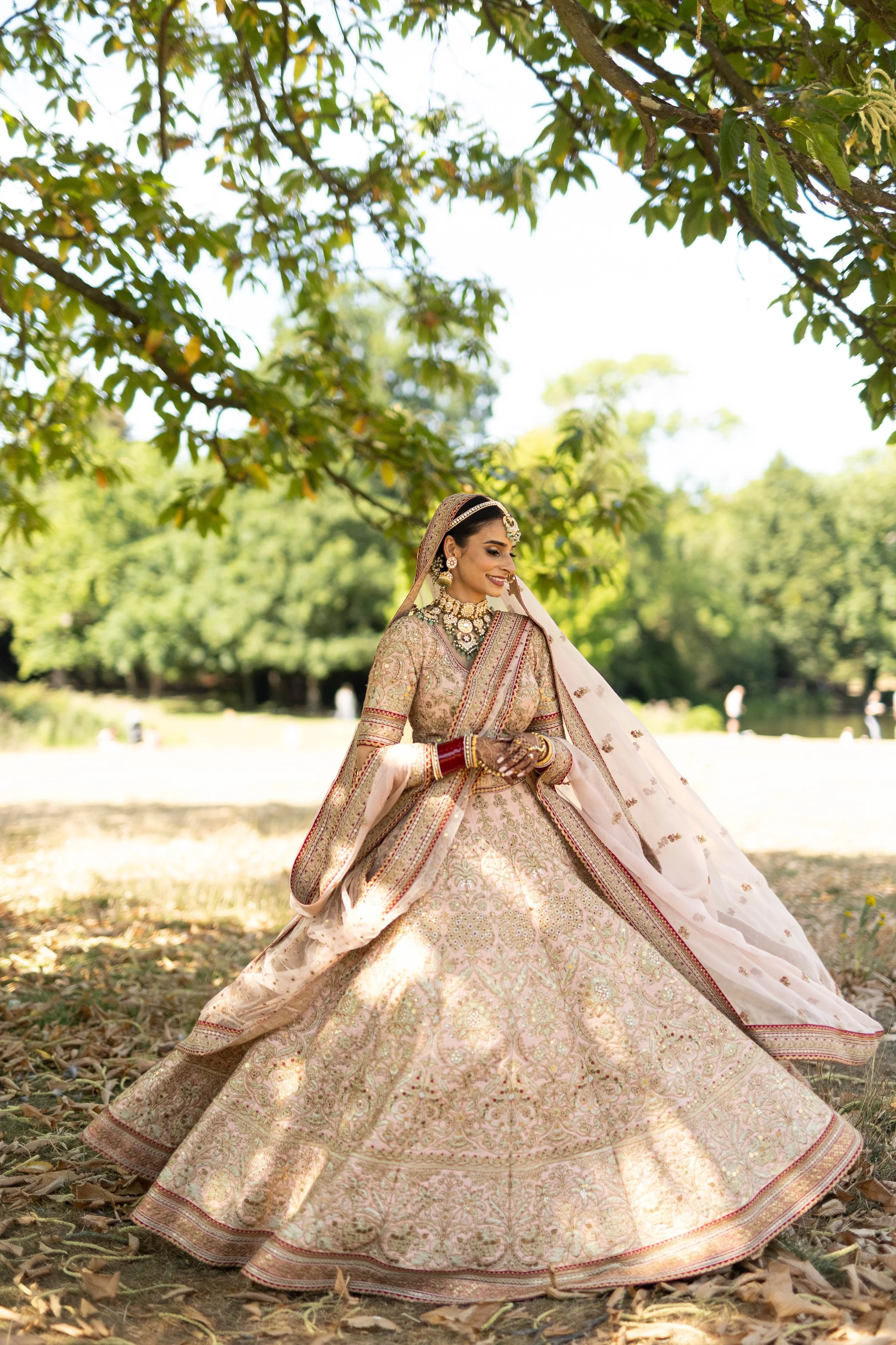 A woman dressed in a traditional bridal lehenga with intricate embroidery, standing outdoors under a tree in a serene, sunlit setting.