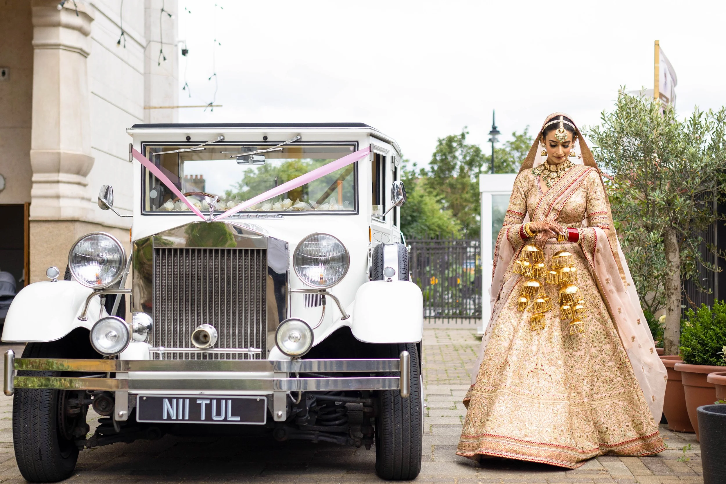 A bride in traditional Indian wedding attire standing beside a vintage white automobile decorated with pink ribbons, indoors with potted plants and a building in the background.