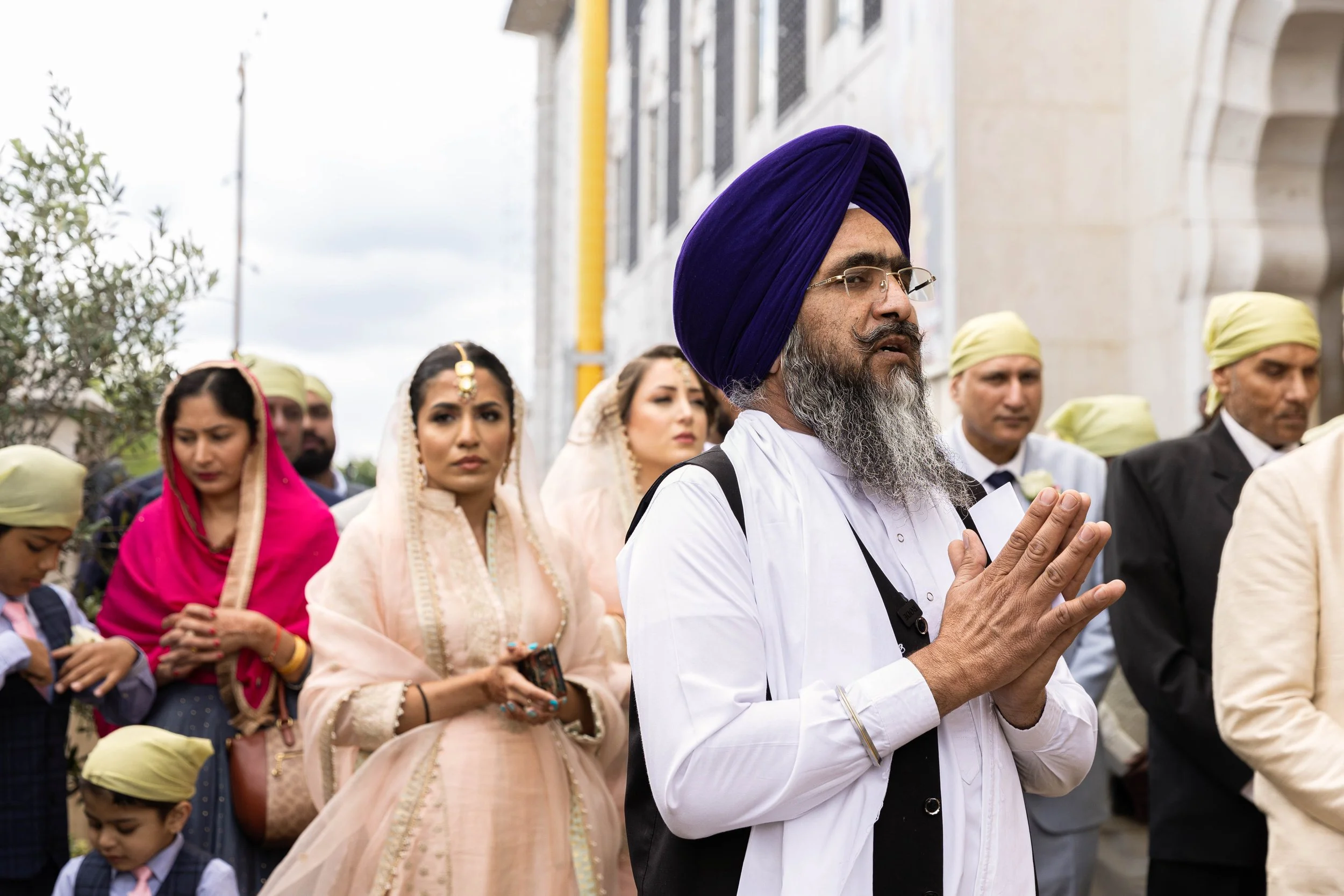 Group of people, including a man with a purple turban and glasses praying with hands pressed together, and women in traditional Indian attire, participating in a religious or cultural ceremony outdoors.