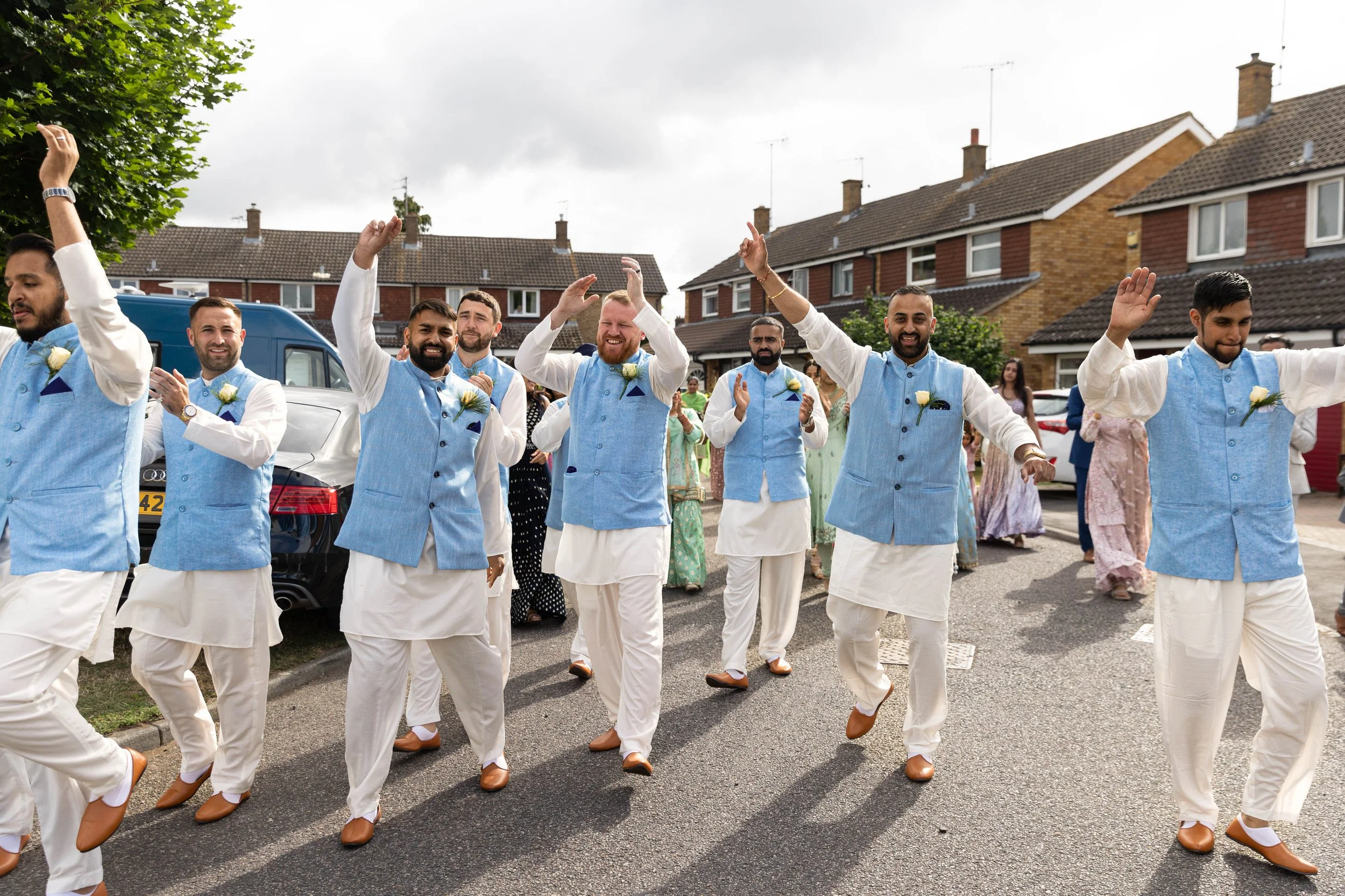 Group of men in traditional South Asian attire dancing in a residential street during a celebration, with women and onlookers in the background.