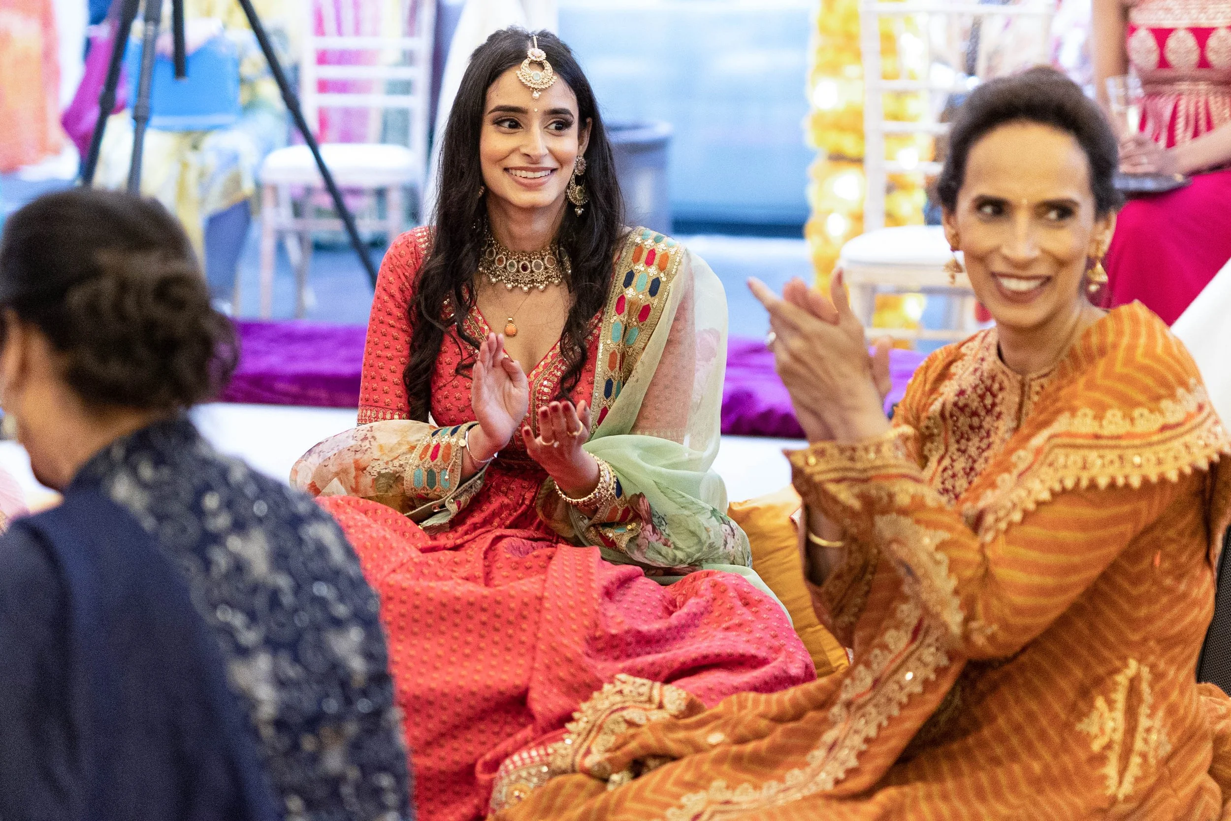 Women dressed in colorful traditional Indian attire at a festive celebration, sitting on the floor and smiling.