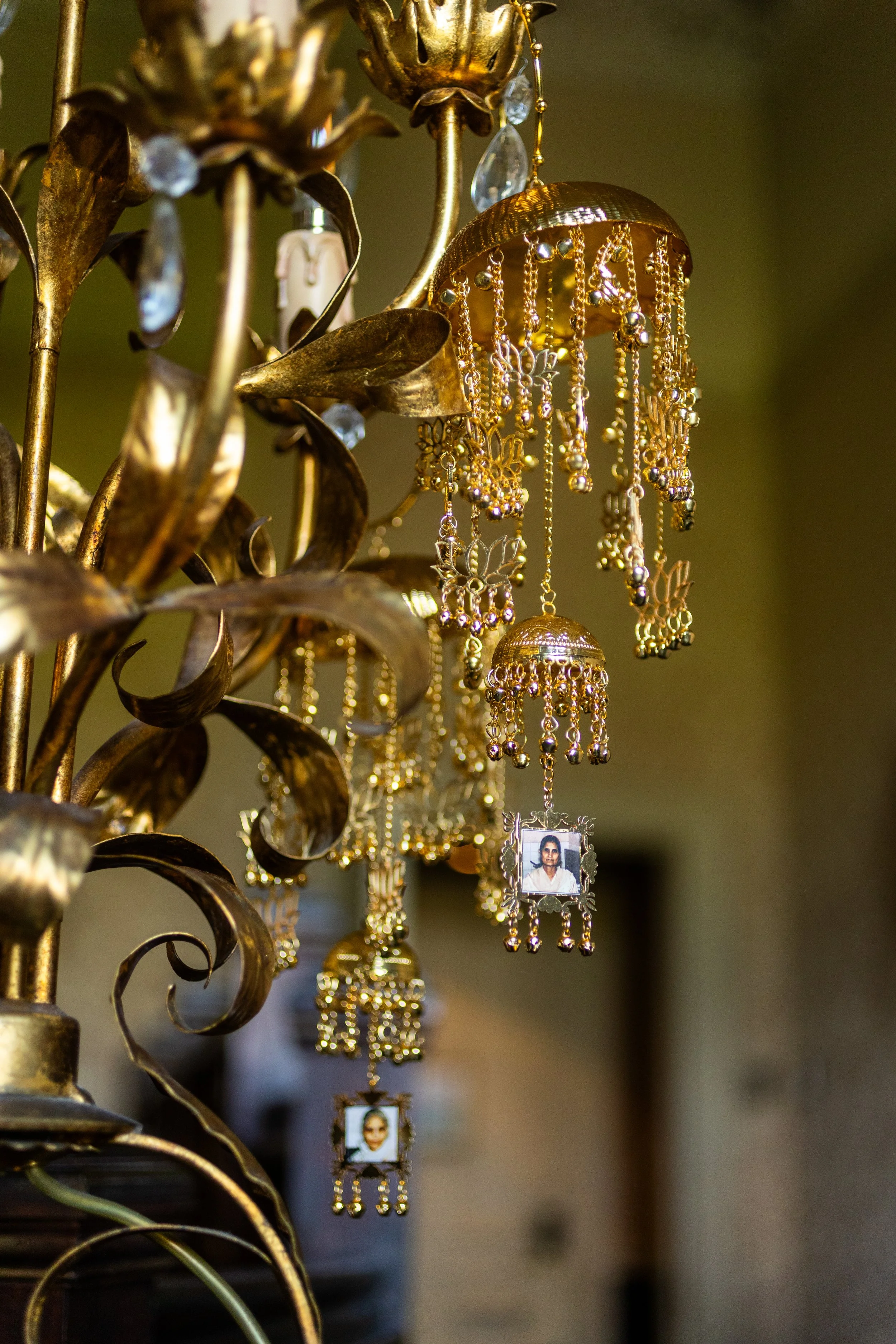 Close-up of a gold decorative chandelier with hanging photo frames and jewelry-like embellishments, ornate metallic leaves, and flowers, with a blurred indoor background.