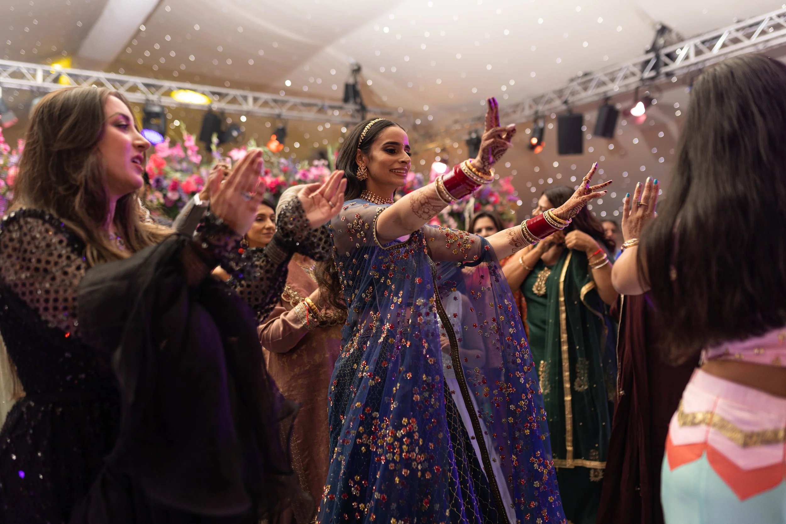 Women dressed in traditional Indian attire dancing and celebrating at a festive event, with floral decorations and string lights overhead.