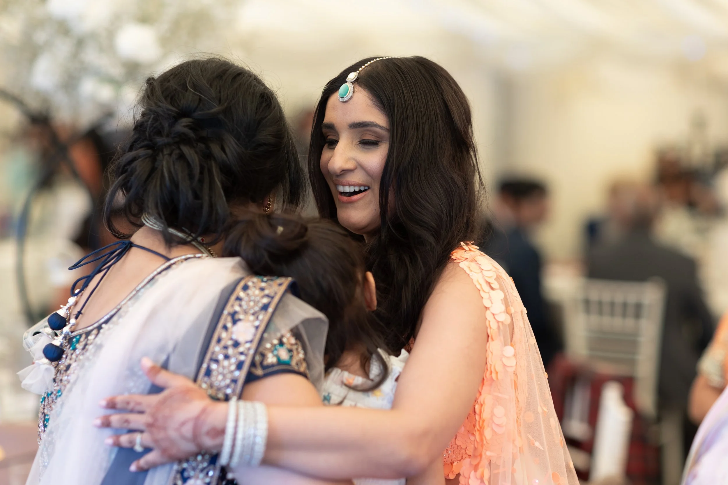 Three women, two in traditional Indian attire and jewelry, sharing an emotional and joyful embrace at a celebration or wedding in a decorated indoor venue.