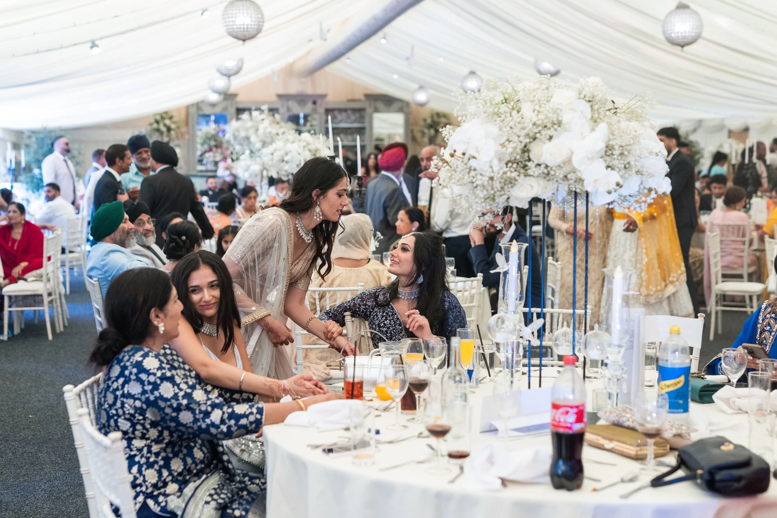 Guests at a wedding reception sitting and socializing at decorated tables under a white tent with floral centerpieces.