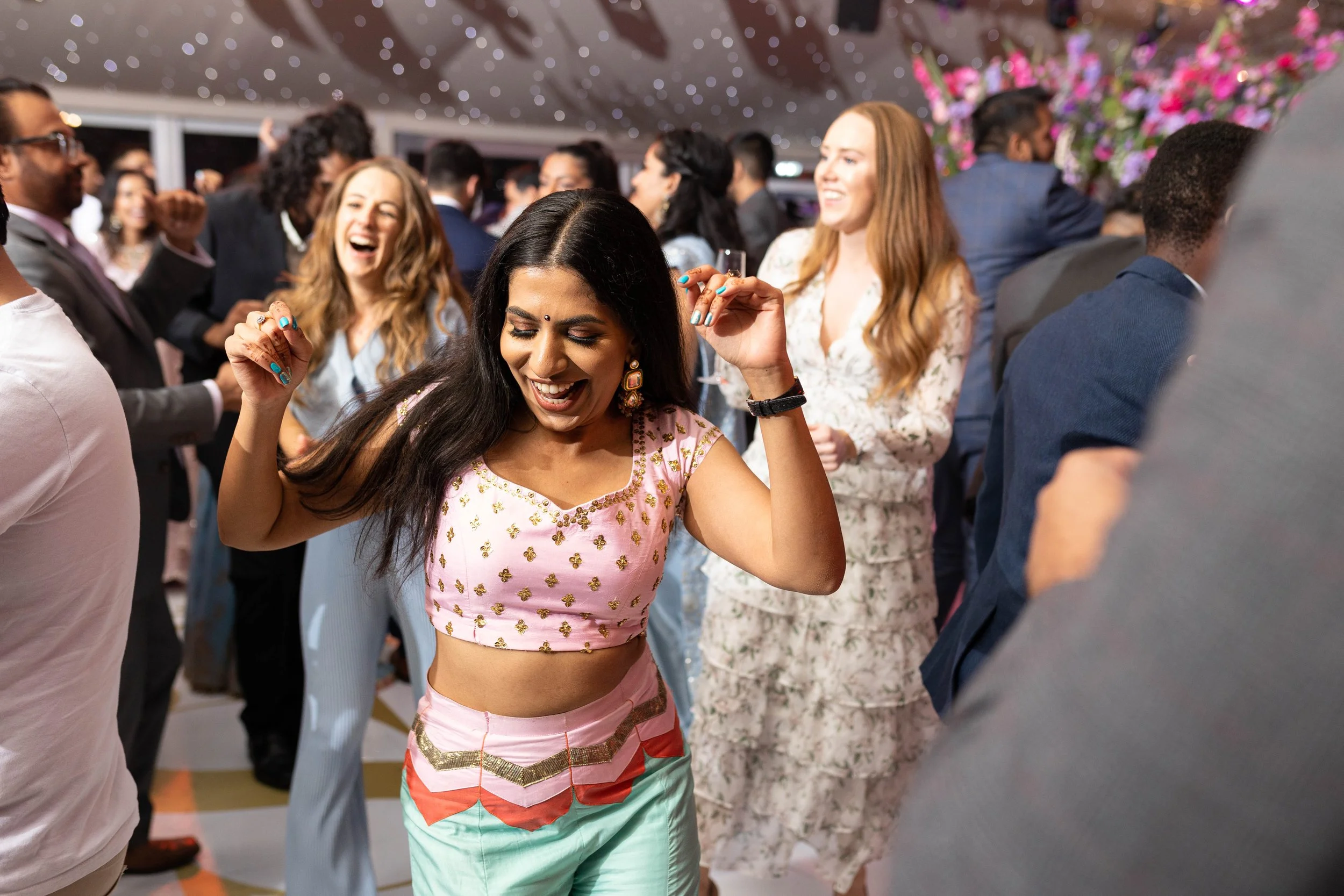 A diverse group of people dancing and enjoying themselves at a lively indoor celebration or party, with a woman in the foreground dancing in a pink and green outfit, smiling and laughing.