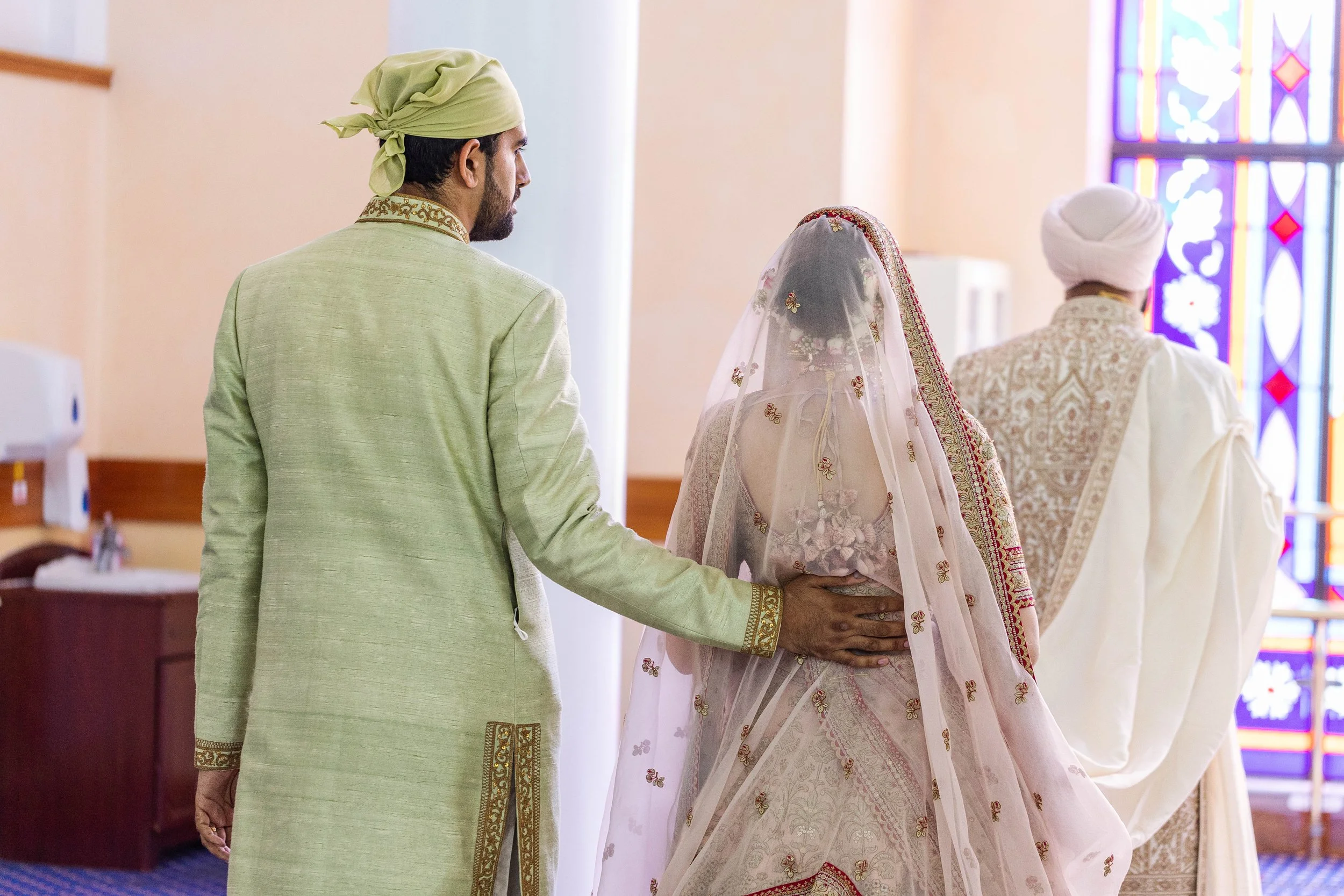 Indian wedding ceremony with bride in traditional white and gold sari and groom in green sherwani, with priest in white robes and turban, in front of stained glass windows.