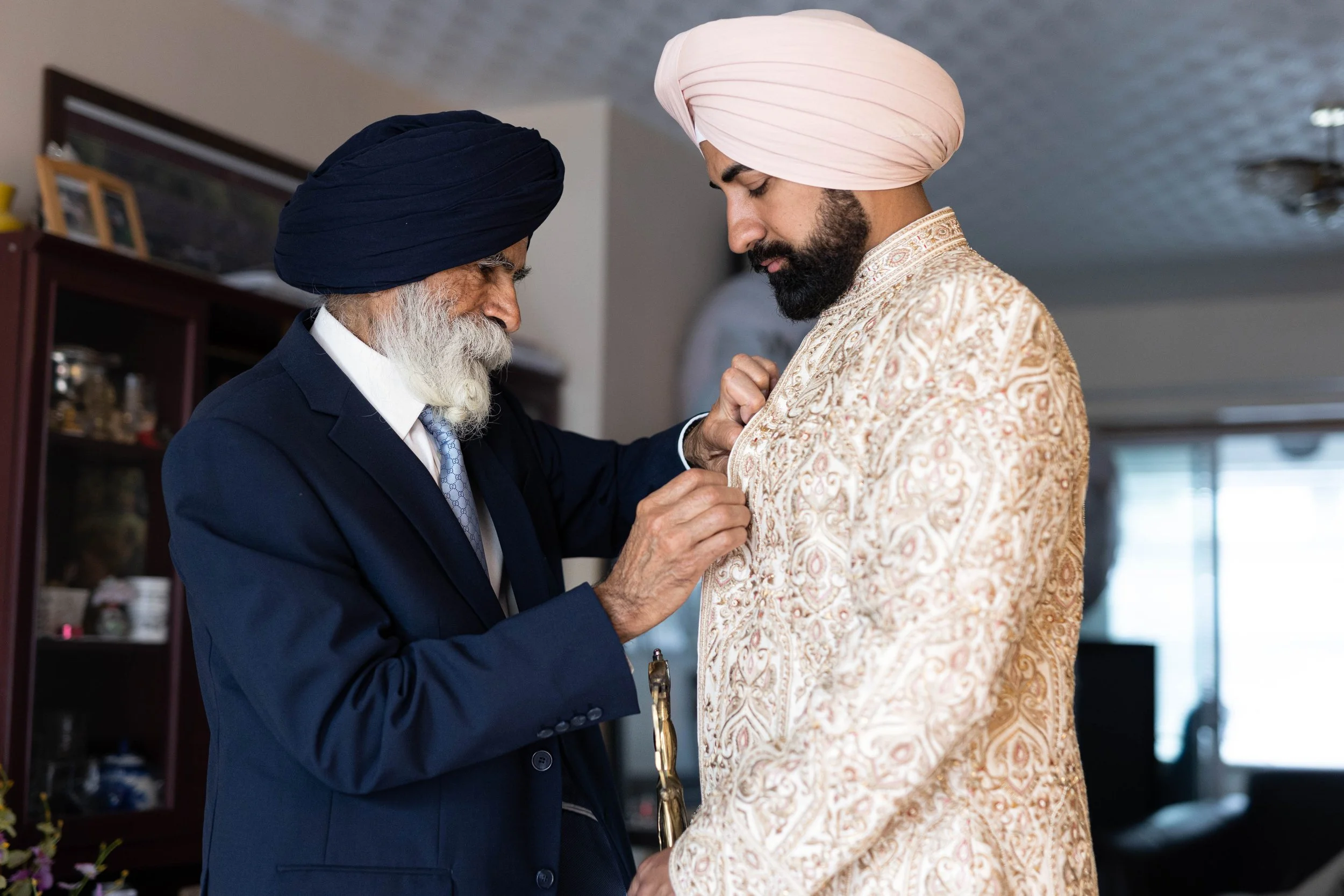 An elderly Sikh man in a dark suit adjusts the turban of a younger Sikh man wearing a light-colored, ornate traditional sherwani during a wedding or formal ceremony.