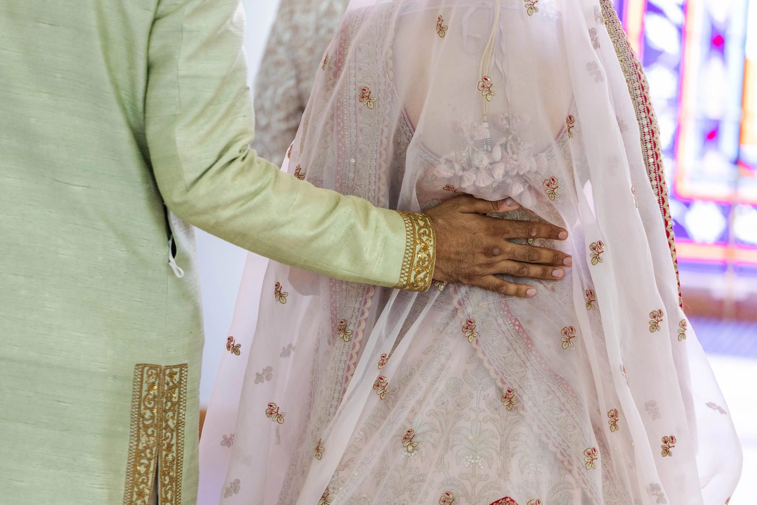 Close-up of a groom's hand resting on a bride's waist, both dressed in traditional Indian wedding attire, with stained glass window in the background.