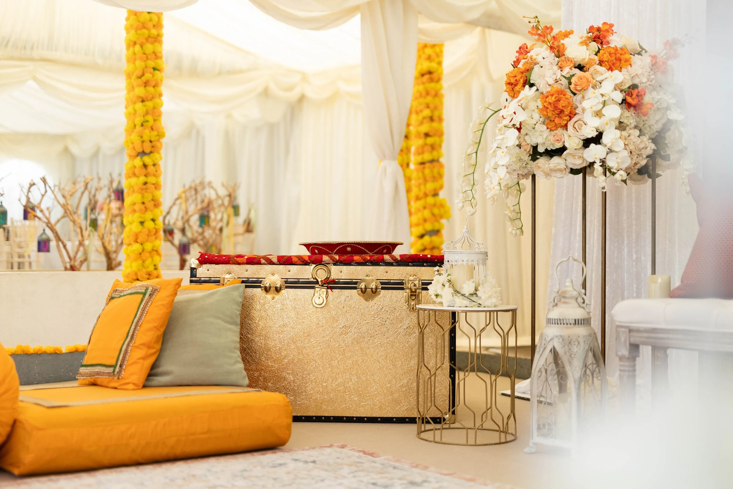 Decorative setup with yellow cushions, a large floral arrangement with white and peach roses and orange flowers in a tall stand, a small white table with lanterns, and draped white curtains at what appears to be a wedding or event venue.