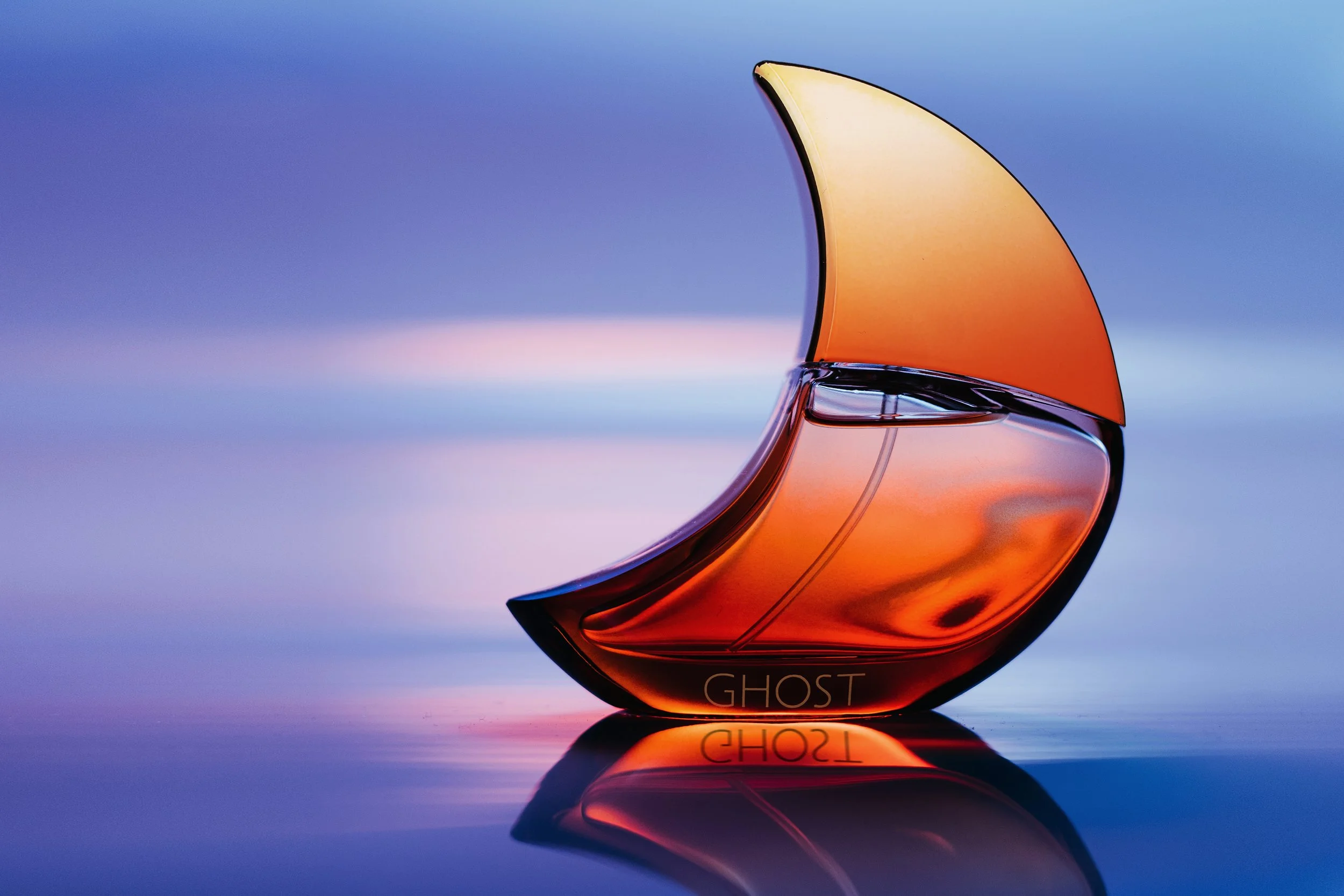 A fragrance perfume bottle shaped like a crescent moon with an orange sail-like lid, labeled 'GHOST', reflecting on a surface with a colorful blurred background