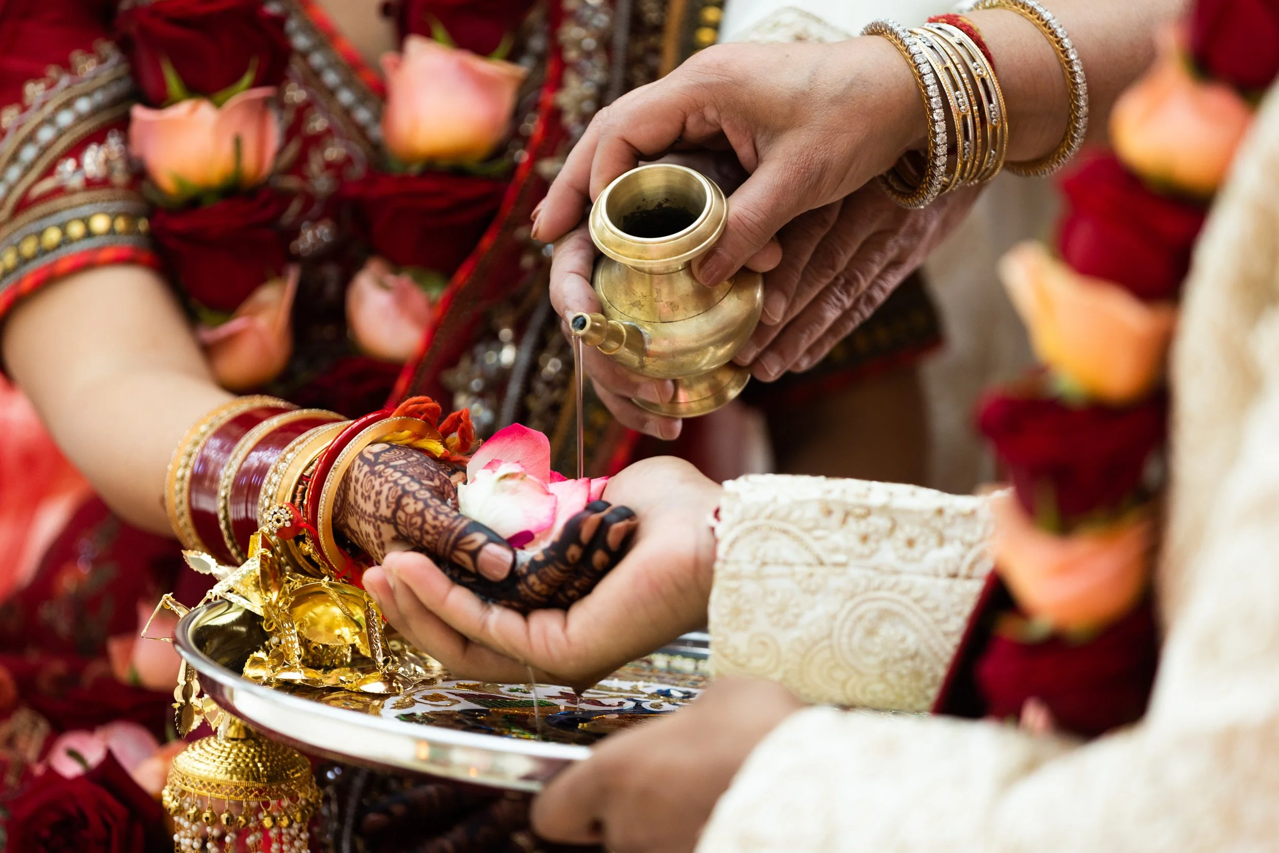 A close-up of a traditional Indian wedding ceremony, showing a groom pouring water over a bride’s hand as part of a ritual, with the bride wearing henna on her hand and traditional jewelry, decorated with rose petals and floral garlands.
