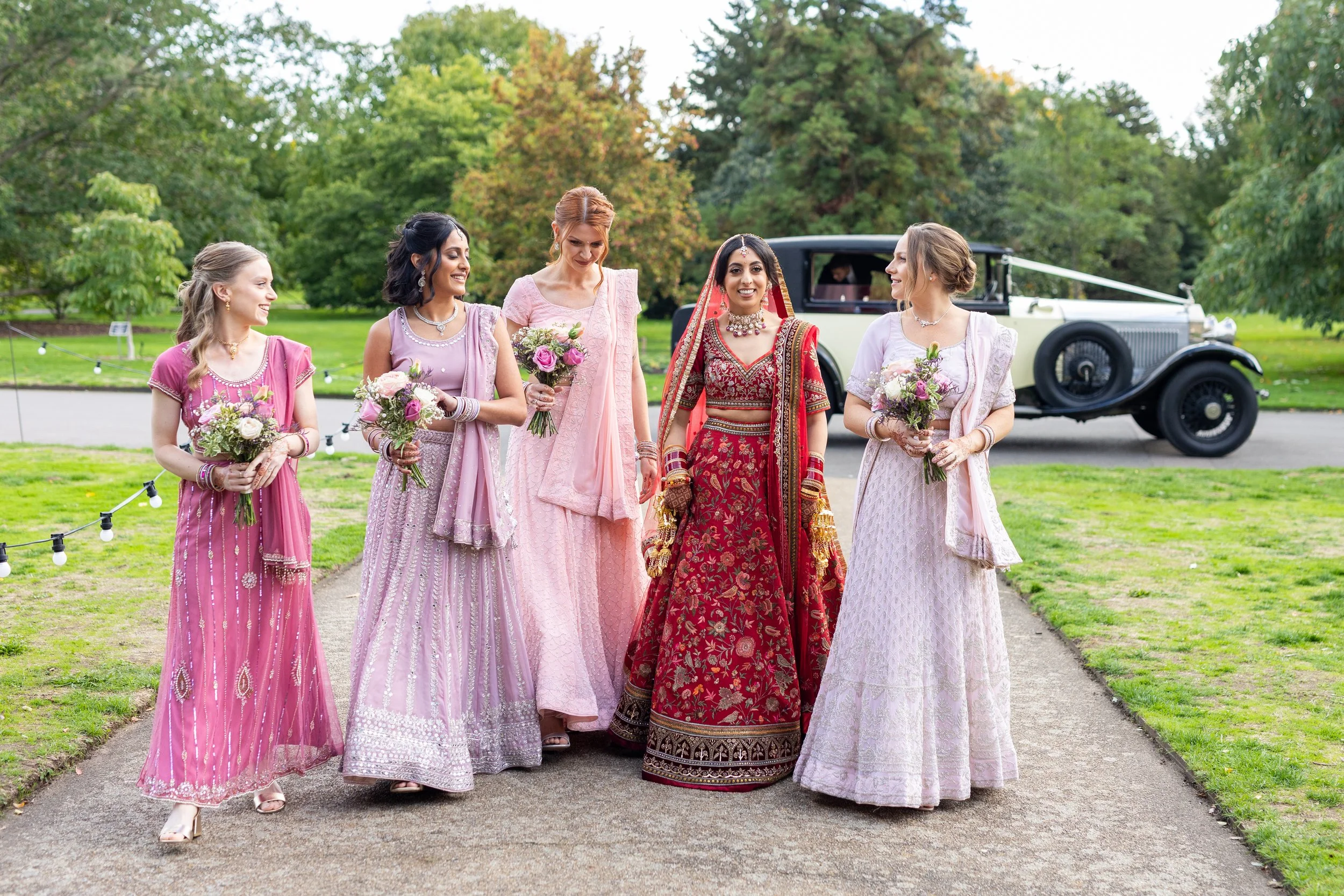 A bride in a red traditional Indian wedding dress walking with her bridesmaids in pink and purple dresses holding bouquets, in a garden with a vintage car in the background.