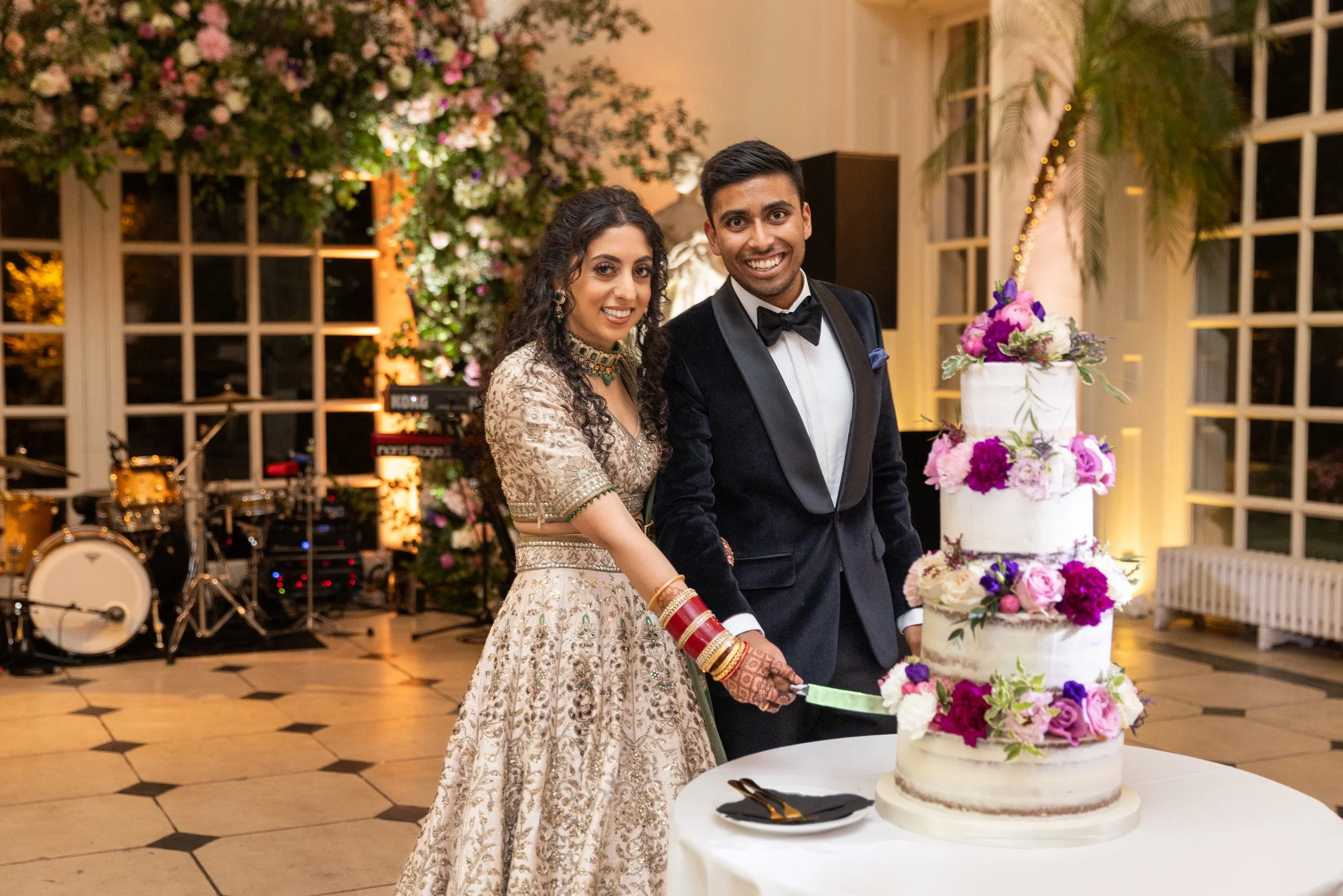 A newlywed couple cutting a wedding cake decorated with pink and purple flowers, in an elegant venue with large windows and floral decorations.