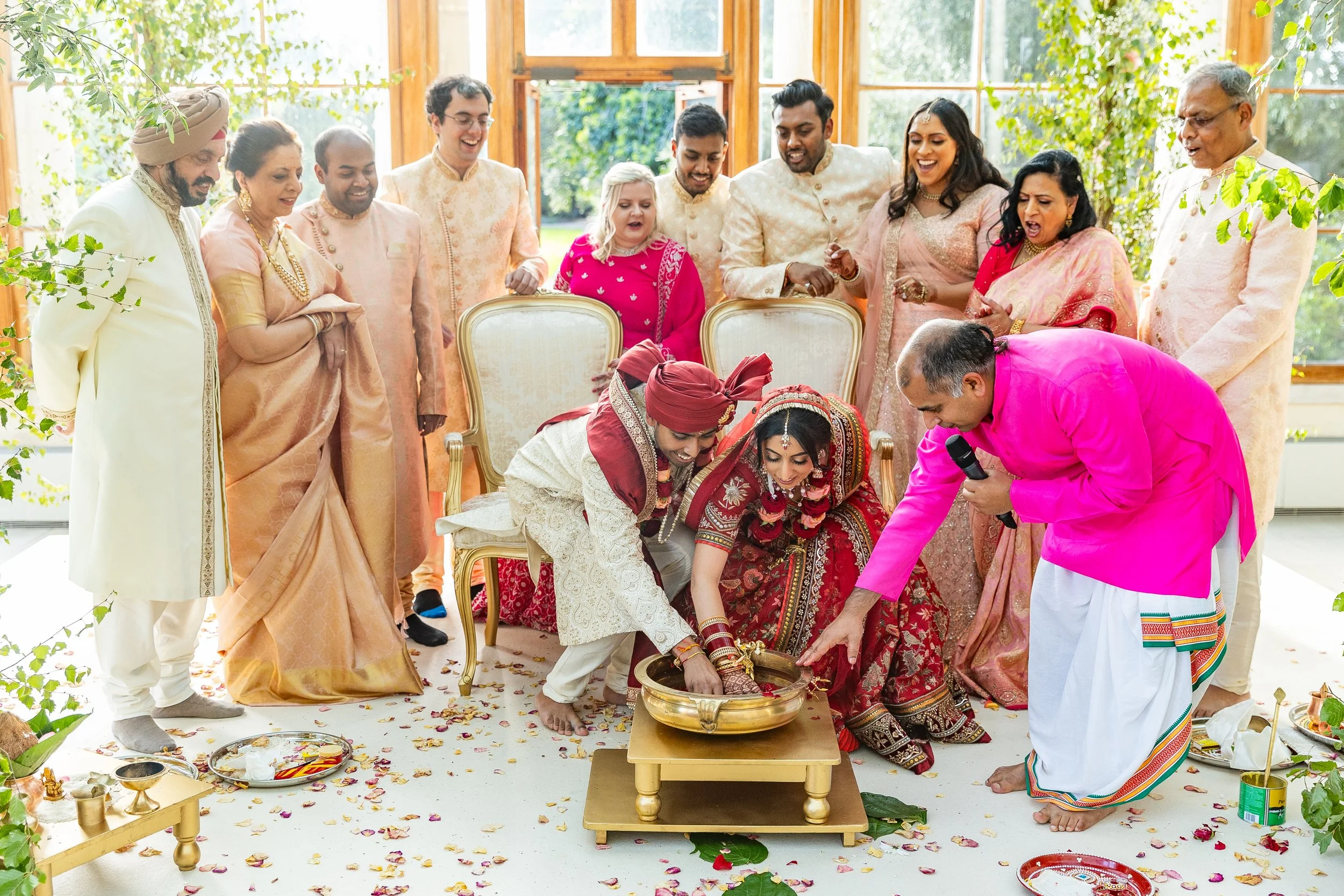 A traditional Indian wedding ceremony with a bride and groom performing rituals, surrounded by family members in colorful attire, inside a bright, flower-adorned venue.