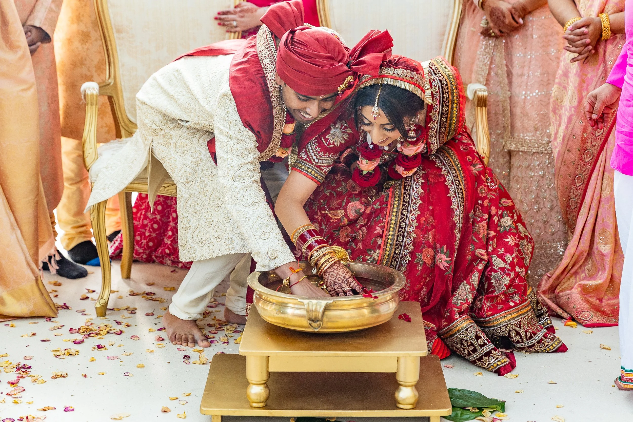 Indian bride and groom performing a traditional wedding ritual, touching water in a gold bowl, surrounded by family members on a decorated wedding stage.
