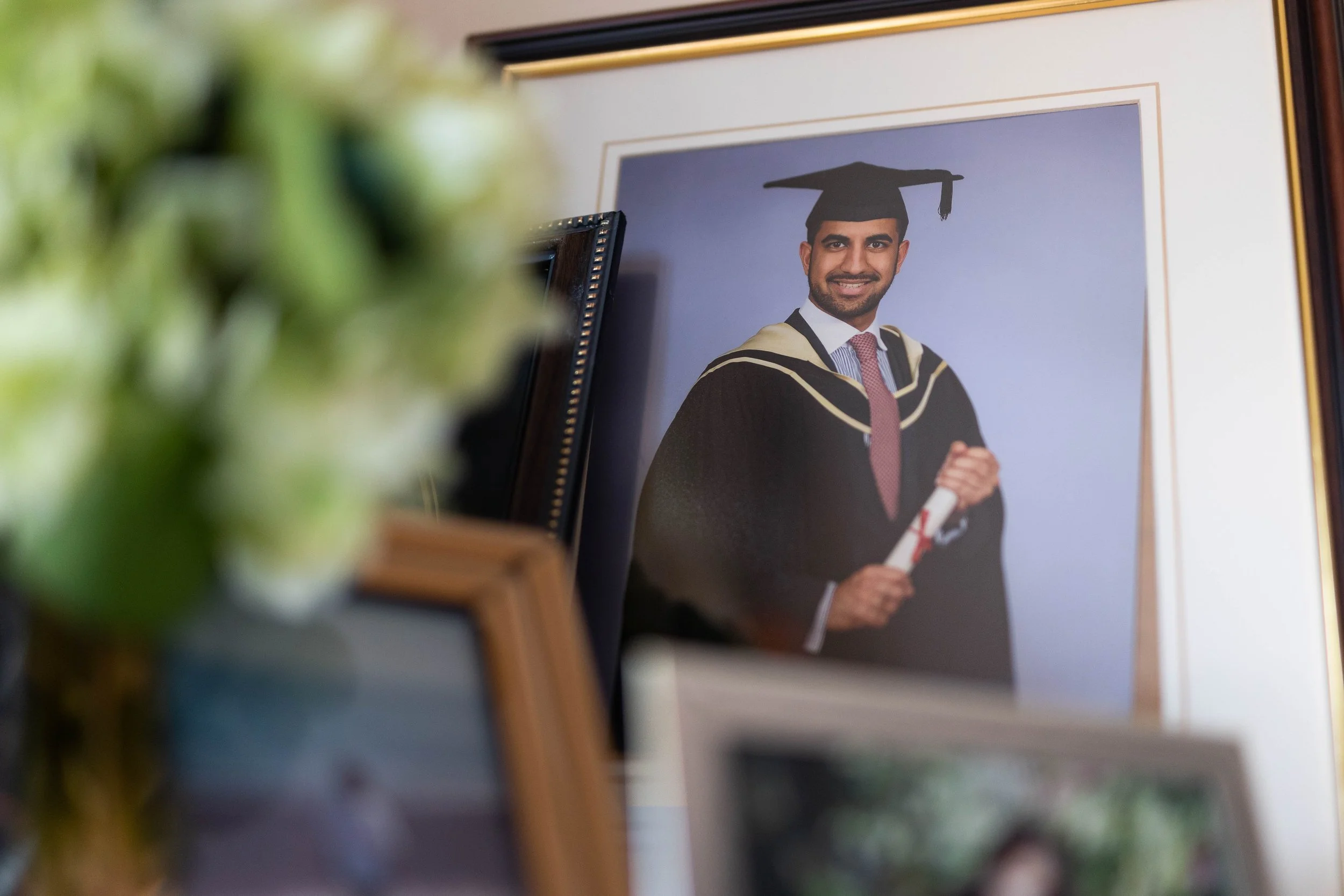 Framed portrait of a man in graduation gown and cap holding a diploma, on a wall. Other framed photos are partially visible in the foreground.