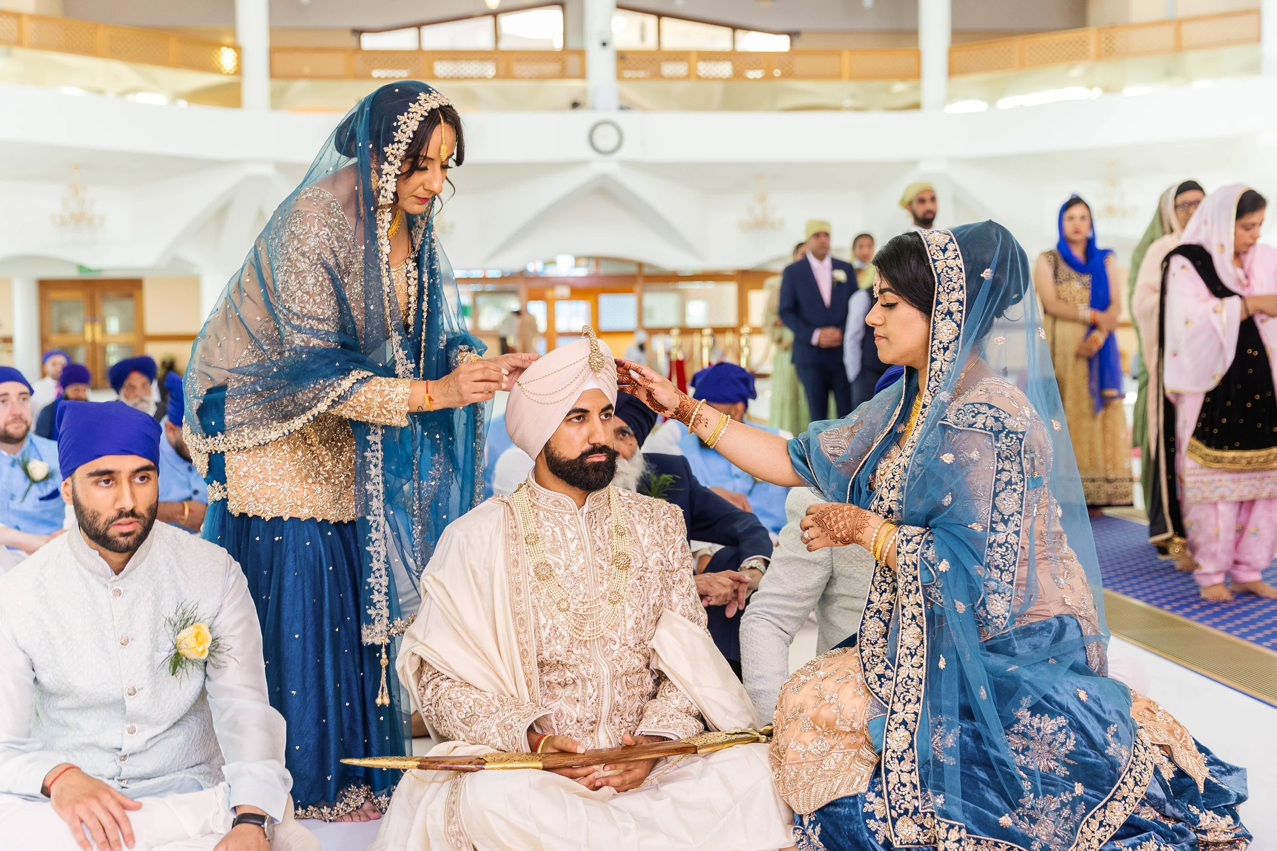 Indian wedding ceremony with bride and groom dressed in traditional attire, surrounded by family and friends in a decorated indoor venue.