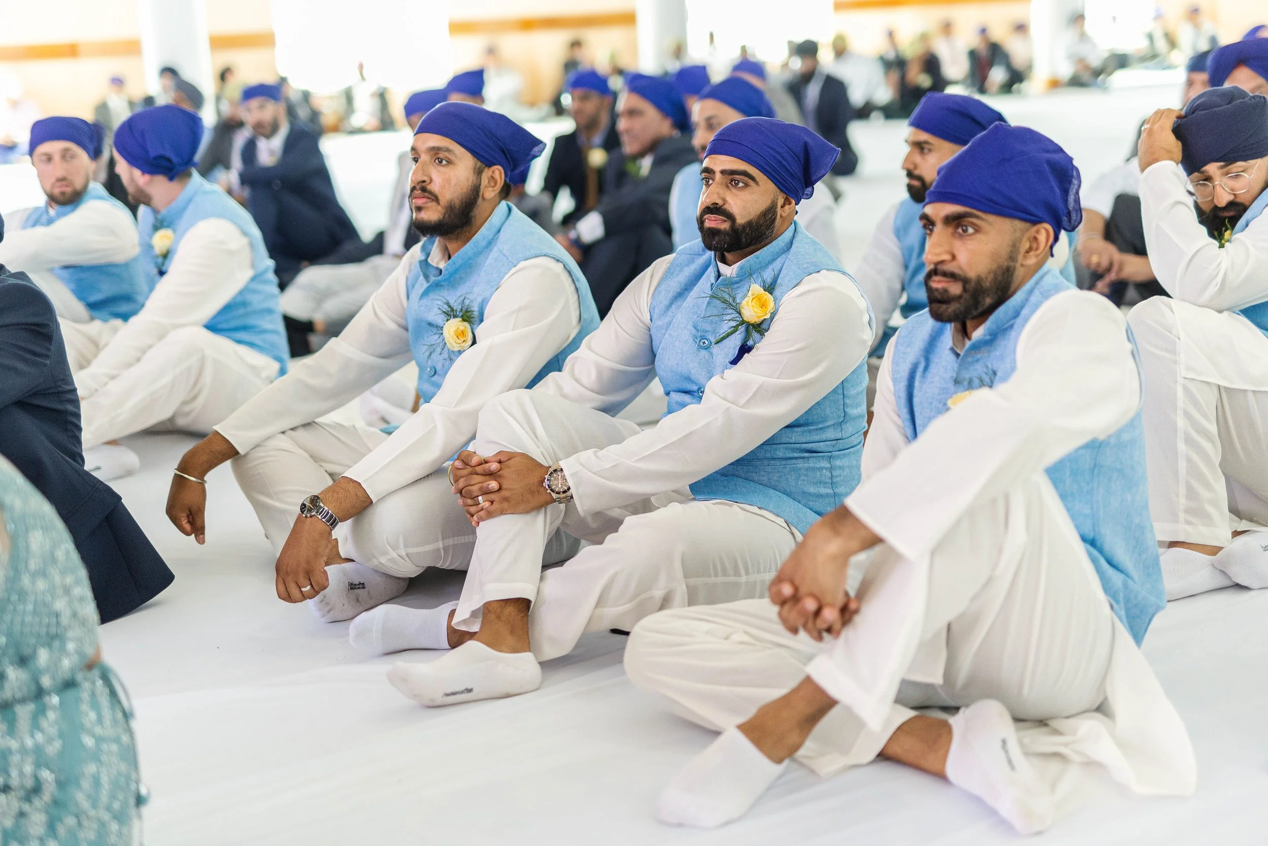 Men dressed in traditional Indian attire sitting on the floor during a ceremony, wearing white kurtas, blue vests, and blue head coverings, with yellow flower boutonnieres.