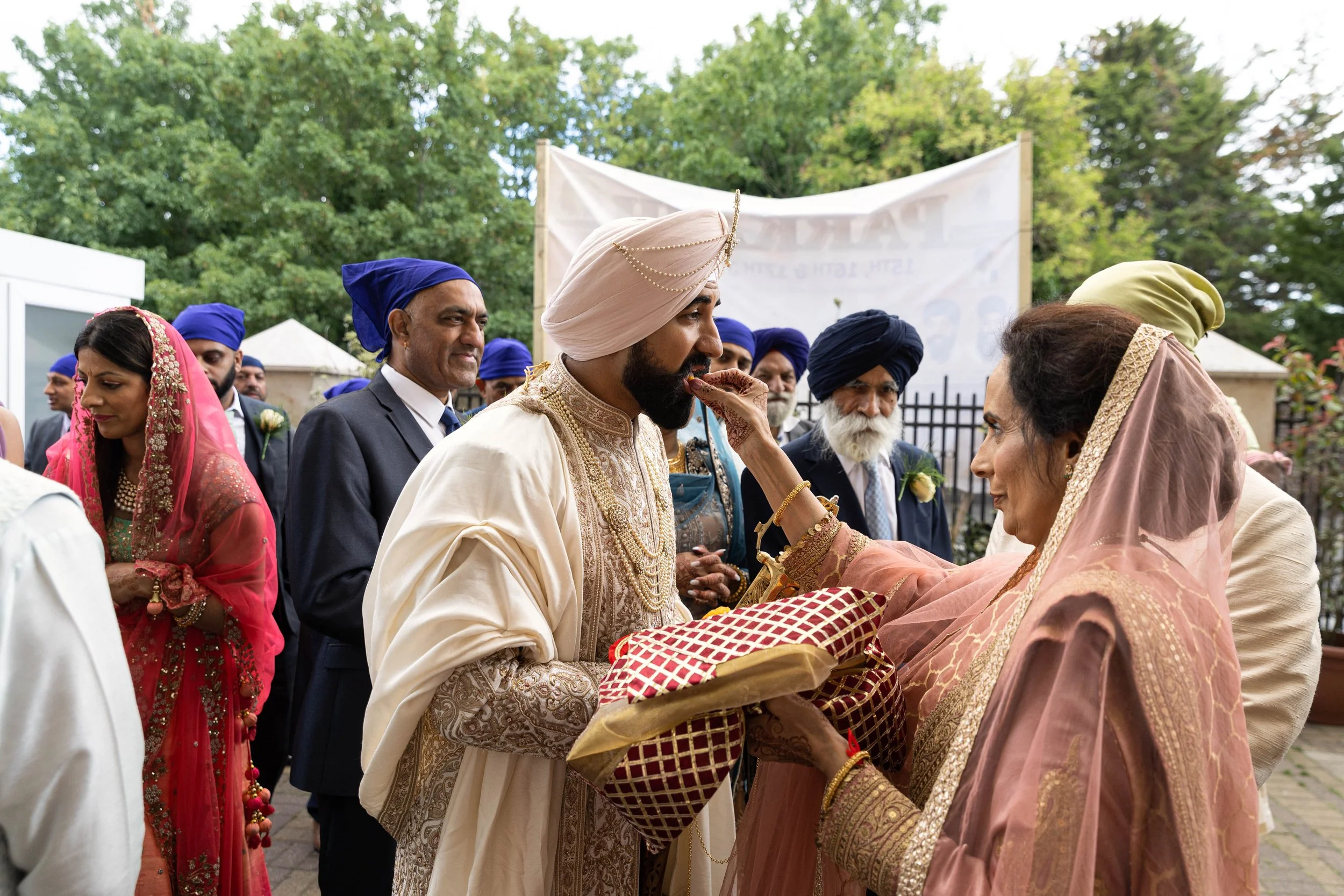 Indian wedding ceremony with groom receiving a traditional blessing from an older woman, surrounded by guests in colorful attire.