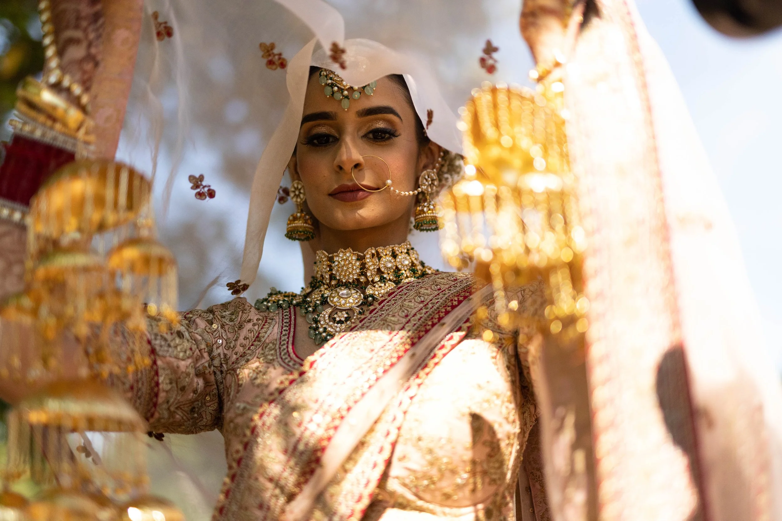 A woman dressed in traditional Indian bridal attire, adorned with intricate jewelry, including a necklace, earrings, and a nose ring, with a garment featuring detailed embroidery and gold accents.