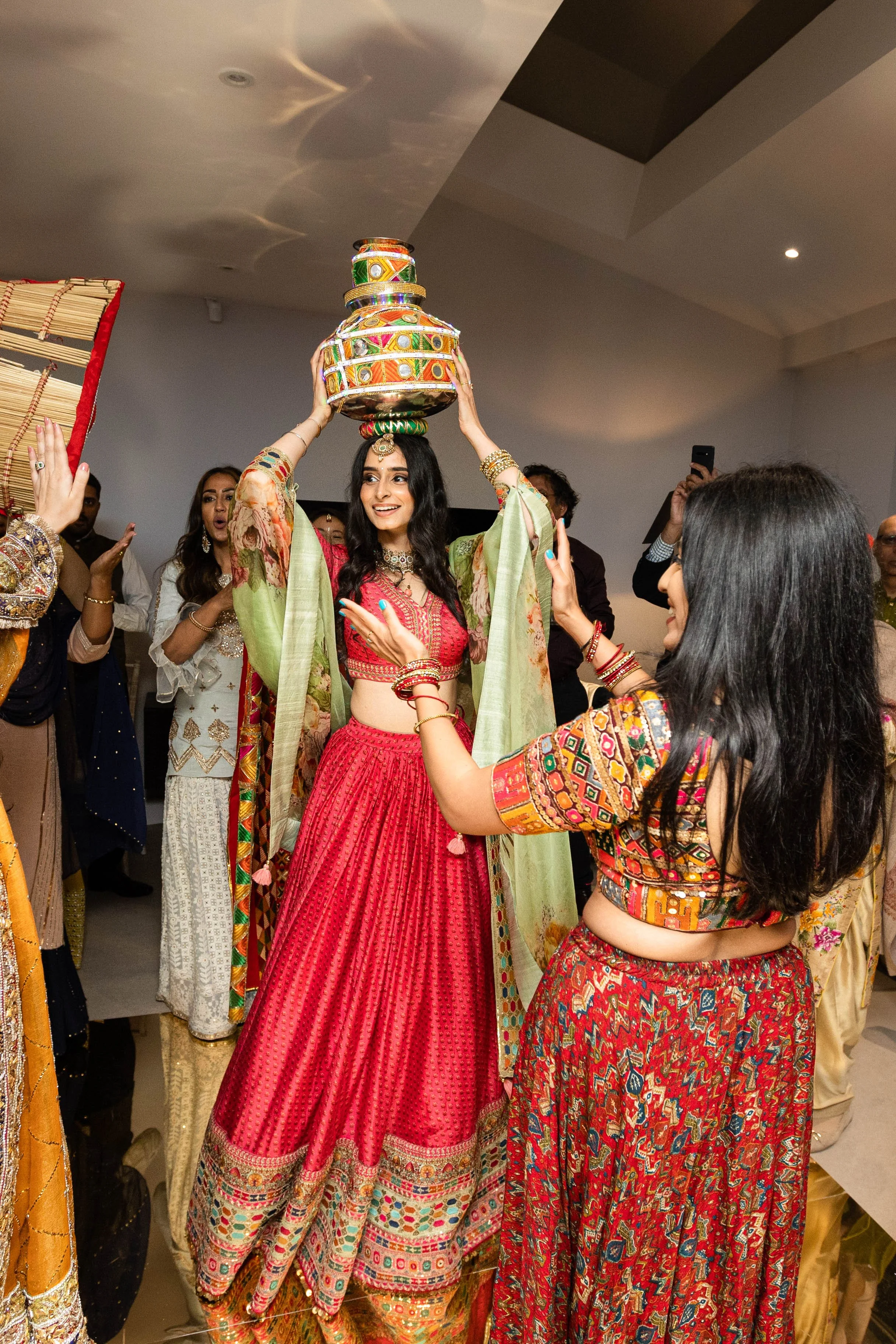 A woman in a red traditional Indian outfit balances a decorated pot on her head during a celebration, surrounded by people dressed in colorful traditional attire.