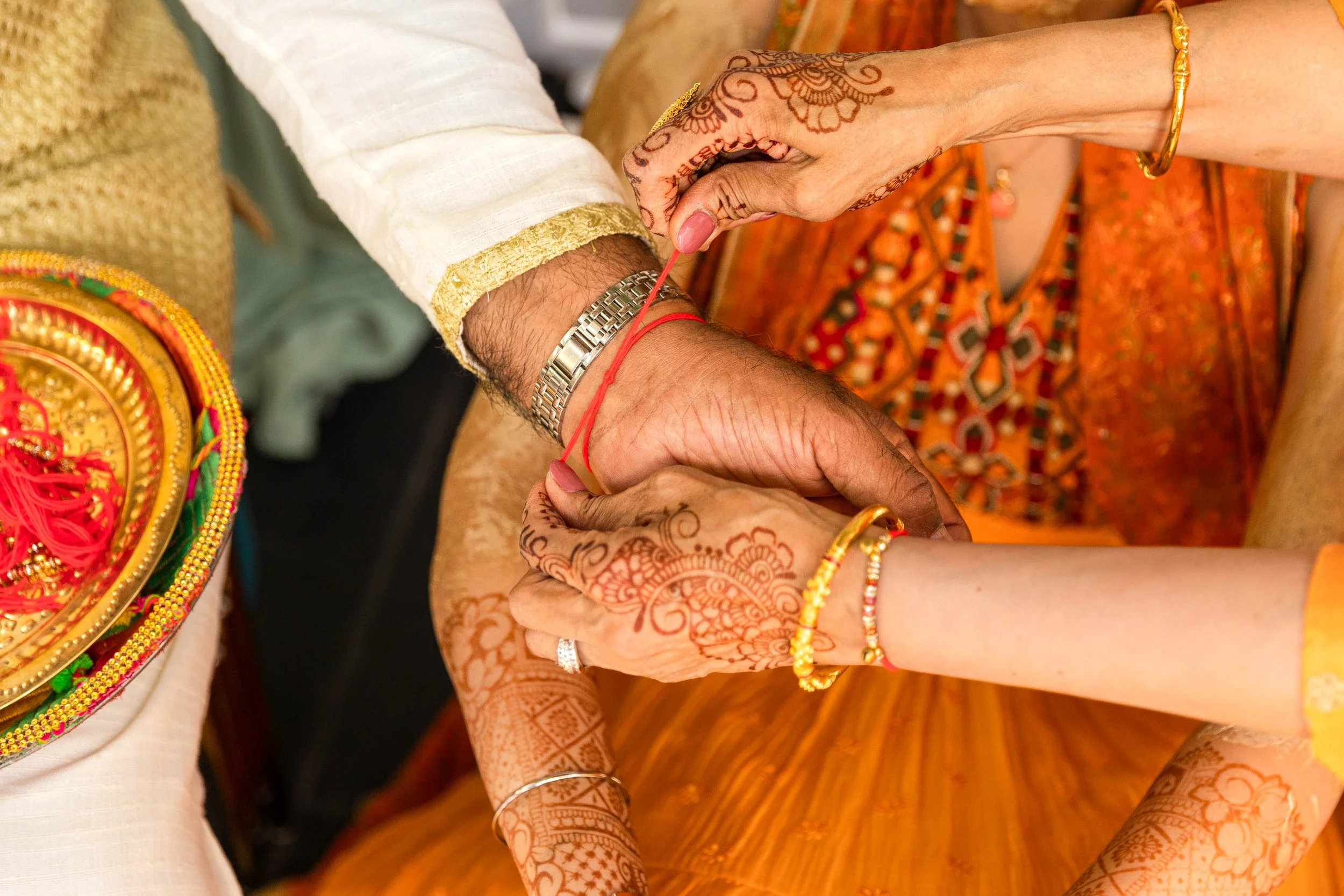 Close-up of an Indian wedding ceremony with the bride and groom holding hands, adorned in traditional colorful attire and intricate henna designs, as the bride applies a red thread to the groom's wrist.
