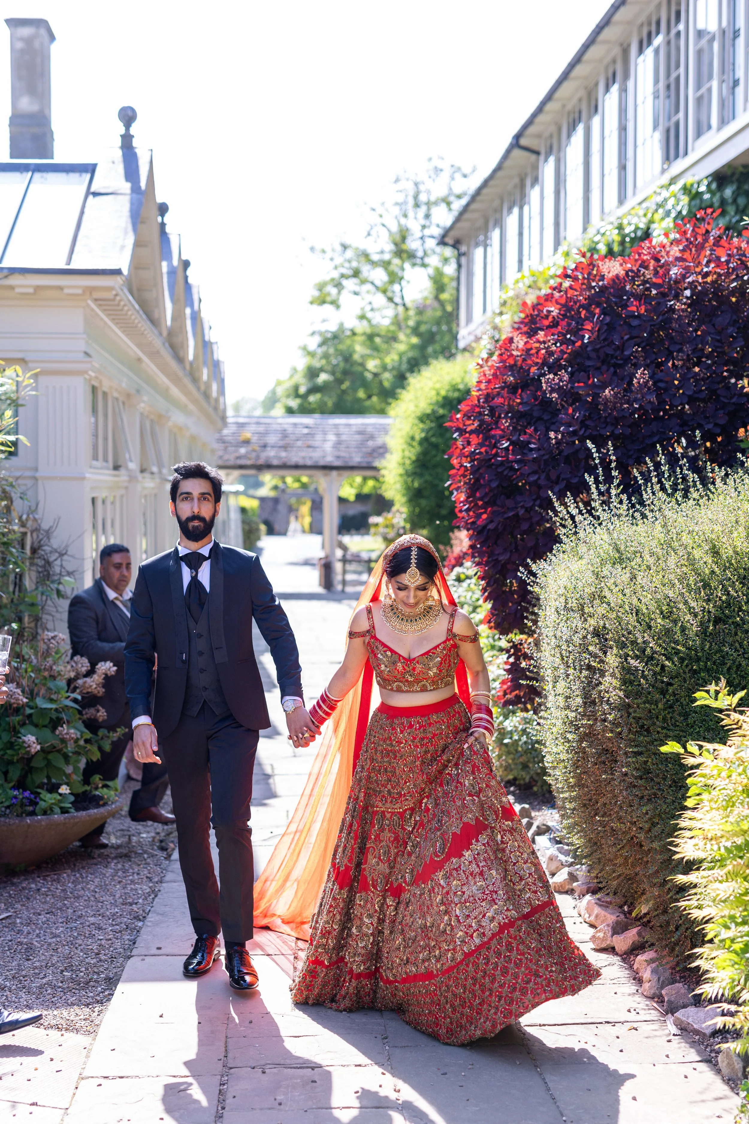 A bride and groom holding hands walking outdoors during a wedding celebration. The bride is dressed in a red and gold traditional Indian wedding attire, and the groom is in a dark suit.