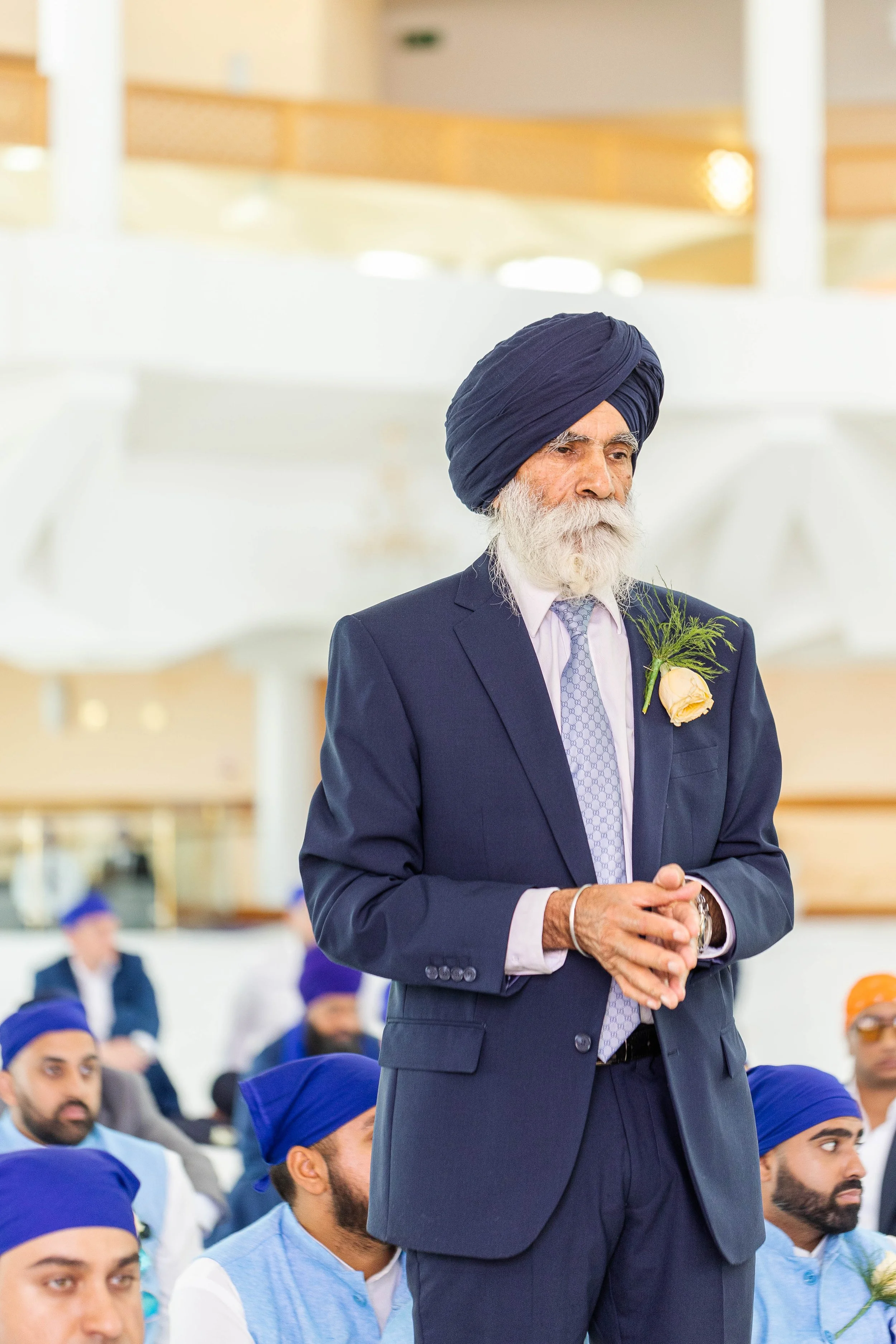 An elderly man with a white beard wearing a dark suit, light tie, and blue turban, standing with hands clasped, during a Sikh gathering.