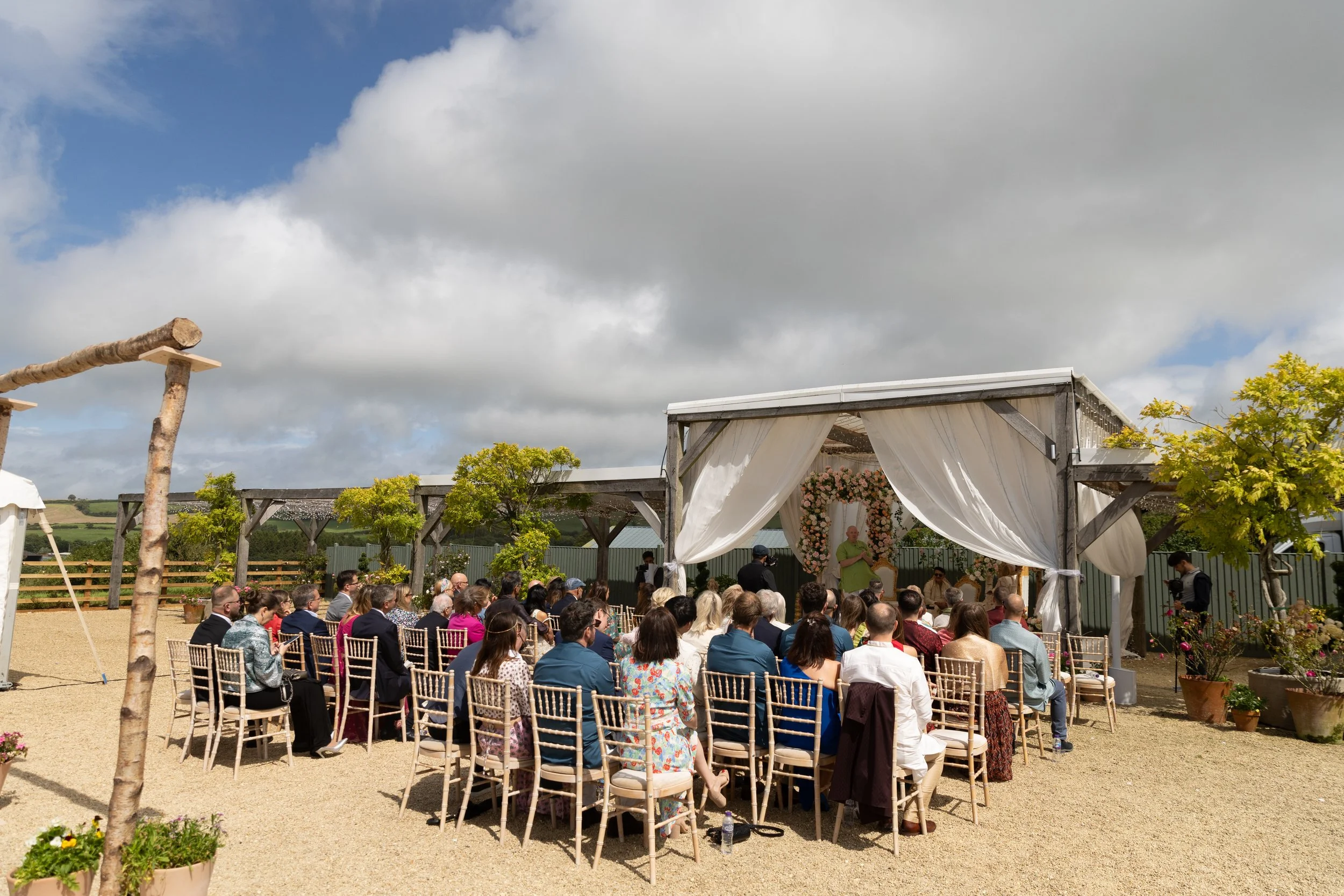 Outdoor wedding ceremony with guests seated under a canopy decorated with white curtains, overlooking a rural landscape with cloudy sky.