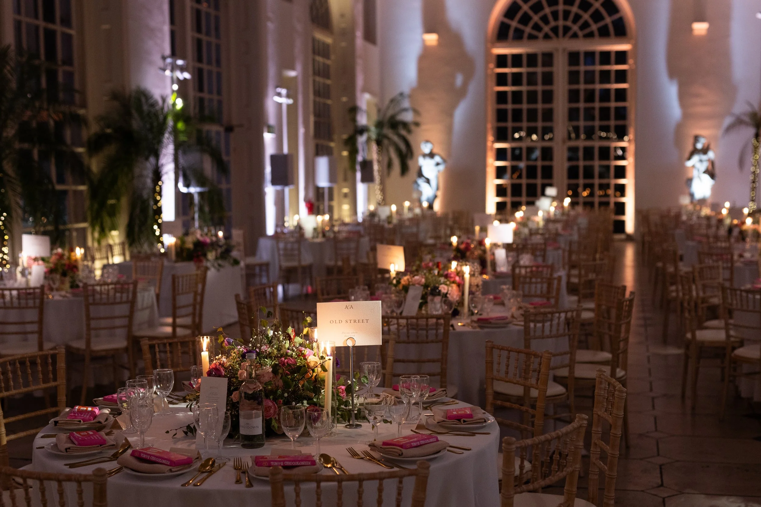 Elegant banquet hall decorated for a wedding reception with round tables, floral centerpieces, candles, and gold utensils, illuminated by soft lighting and large windows.