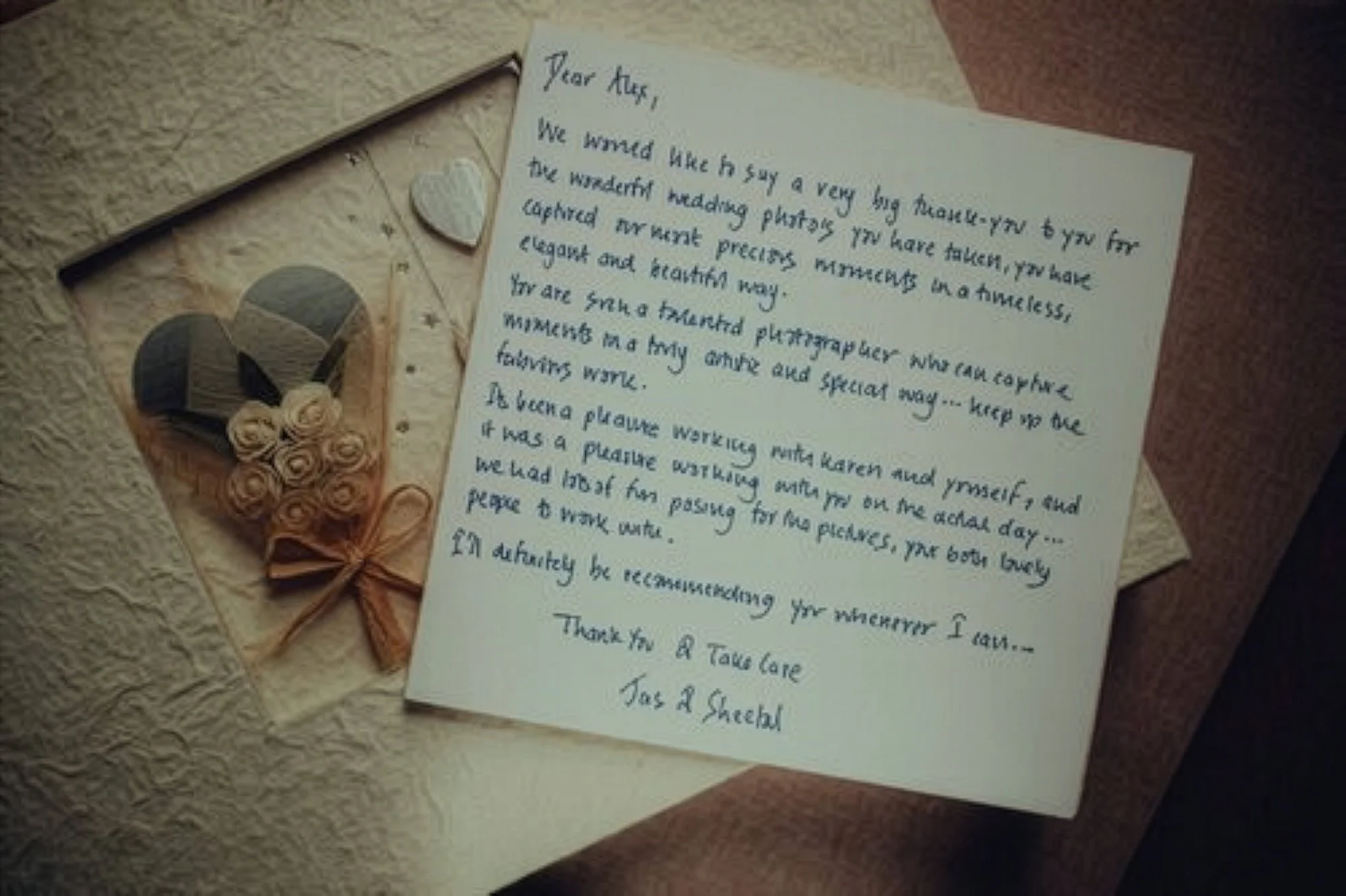 A handwritten thank you note on white paper is placed on a textured surface. Next to it are decorative items, including a heart-shaped wooden piece with small roses attached and a small heart-shaped ornament, tied with a pink ribbon.