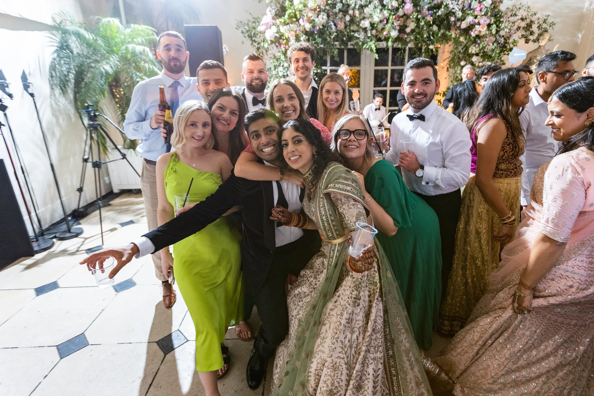 Group of people celebrating at a wedding reception, with some women in traditional Indian attire and men in formal wear, smiling and posing for a photo in a decorated venue with flowers and large windows.