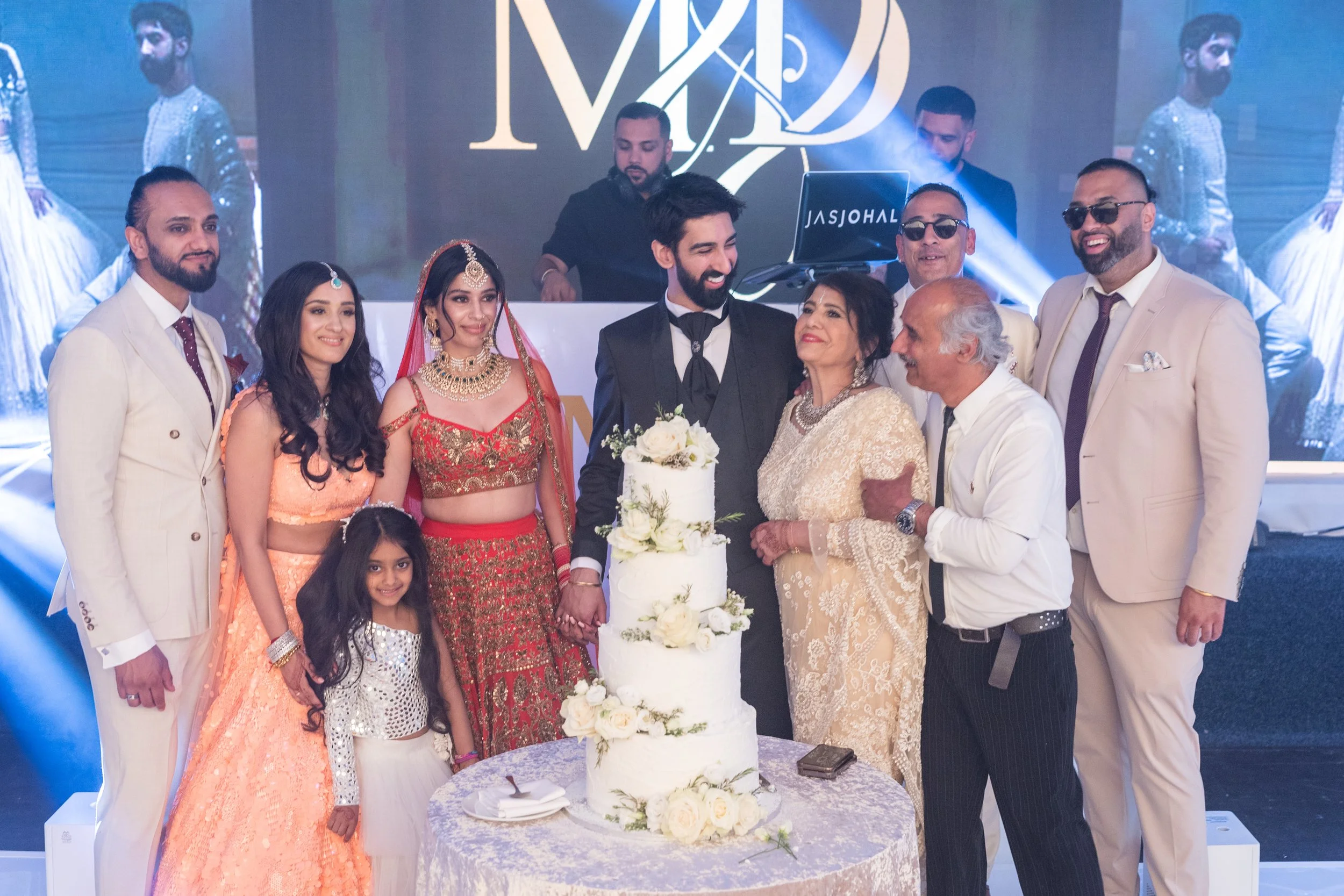 A group of people celebrating a wedding, with the bride and groom in the center, holding hands near a wedding cake. The bride is dressed in traditional Indian attire with jewelry, and the groom is in a black suit. Family and friends surrounding them 
