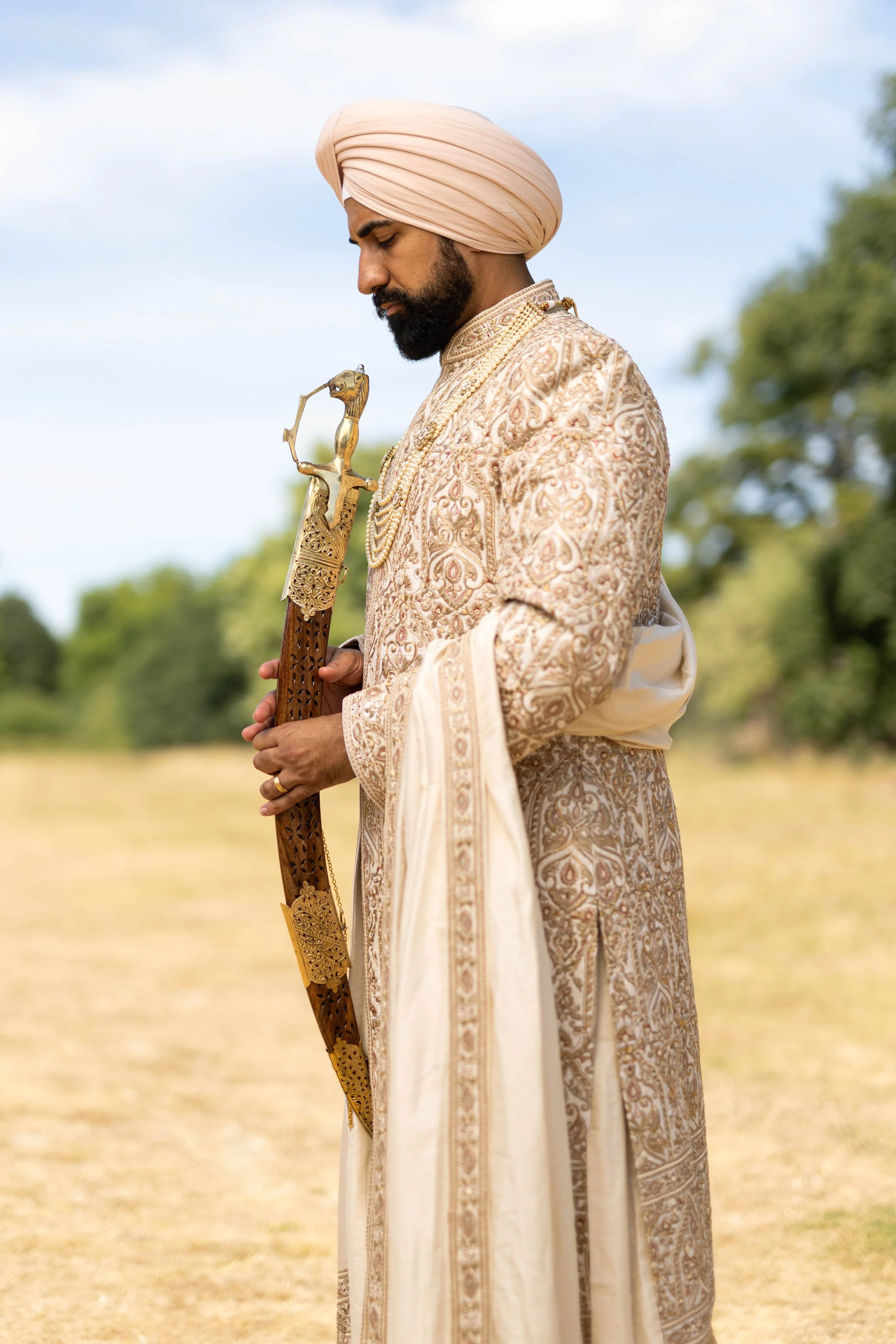 A man dressed in traditional Indian wedding attire, holding a sword, standing outdoors with trees in the background.