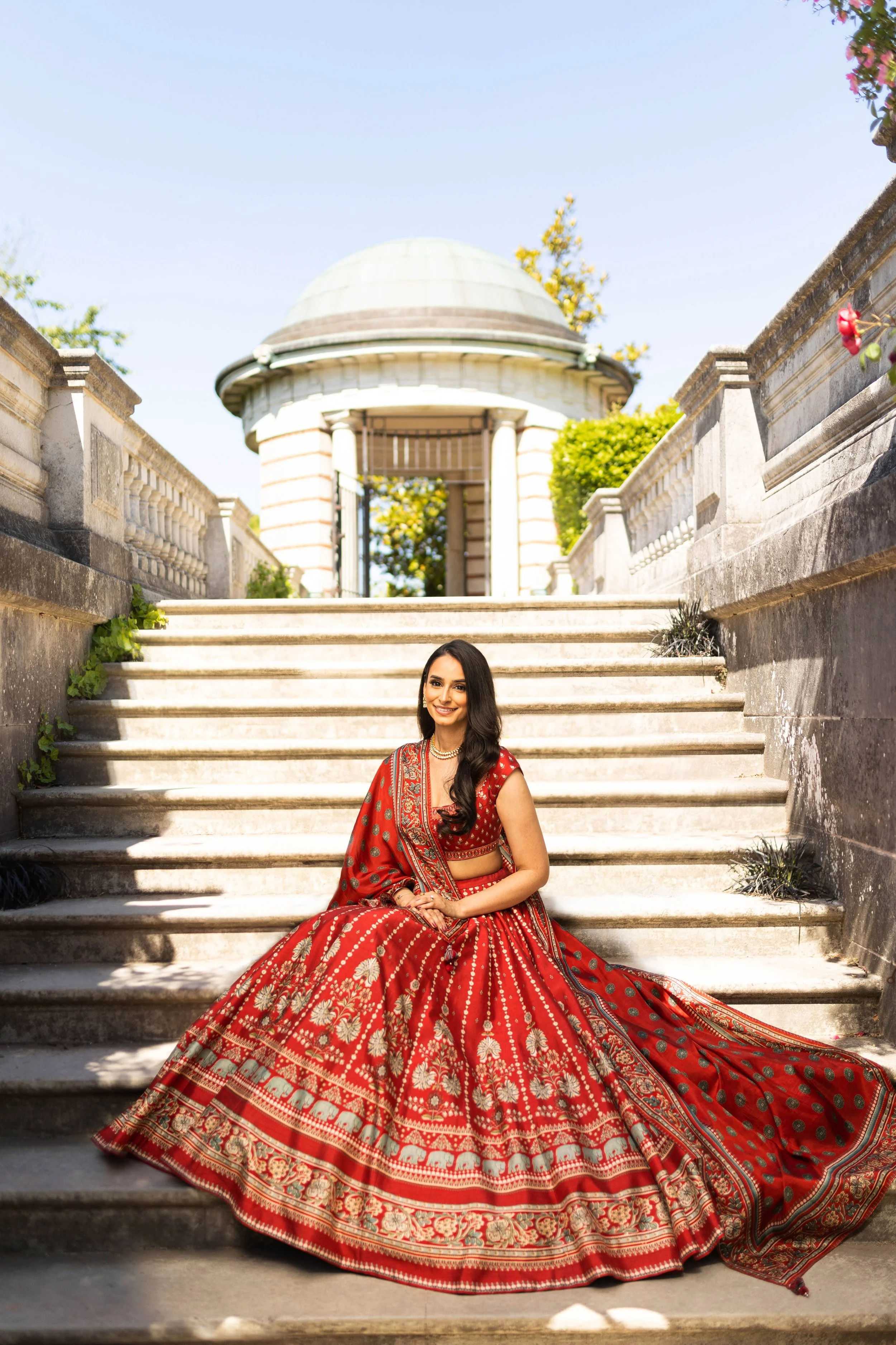 A woman dressed in a traditional red Indian outfit sitting on stone steps outdoors with a historic building in the background.