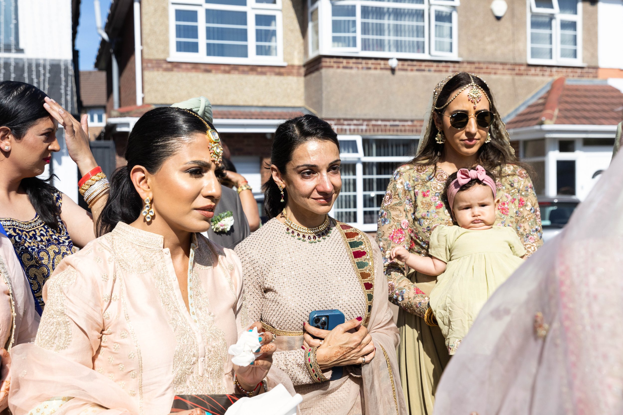 Group of women and a baby at an outdoor celebration or cultural event. The women are dressed in traditional South Asian attire with jewelry, and the baby is wearing a yellow dress with a pink headband. The background shows residential houses.