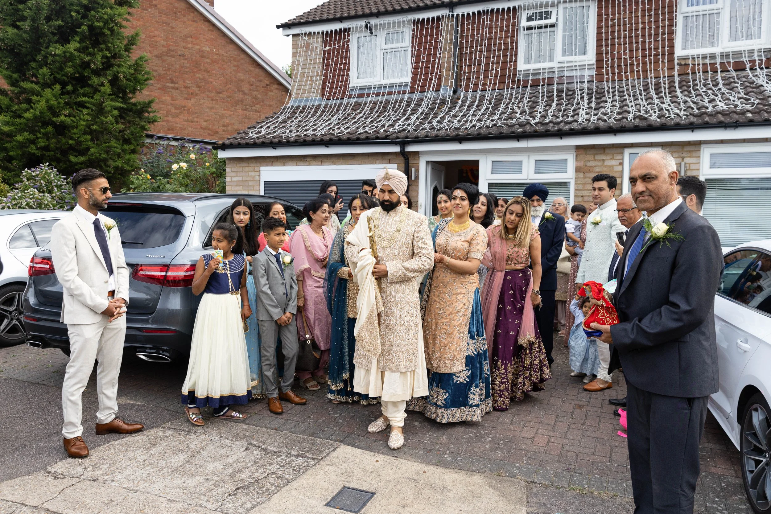A group of people dressed in traditional Indian attire gathered outside a house, some holding flowers and others with hands folded in a prayer or respectful gesture, during what appears to be a festive or ceremonial event.