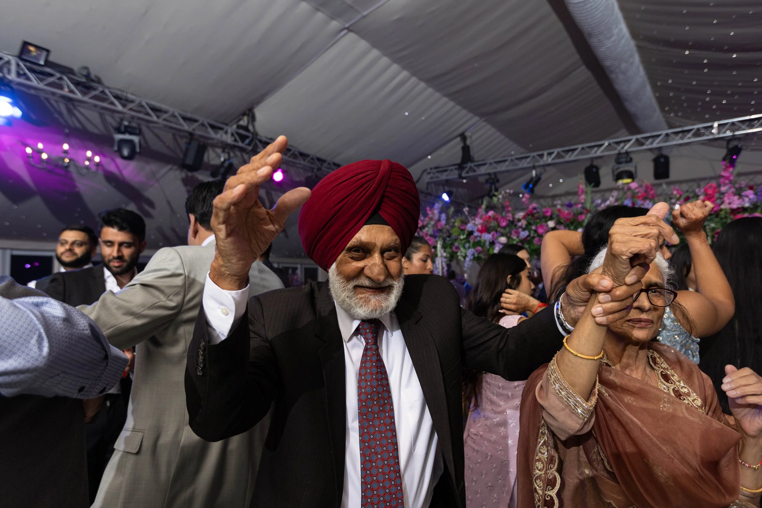 Older man with a red turban dancing with an older woman at a celebration, surrounded by other celebrants in formal attire and floral decorations.