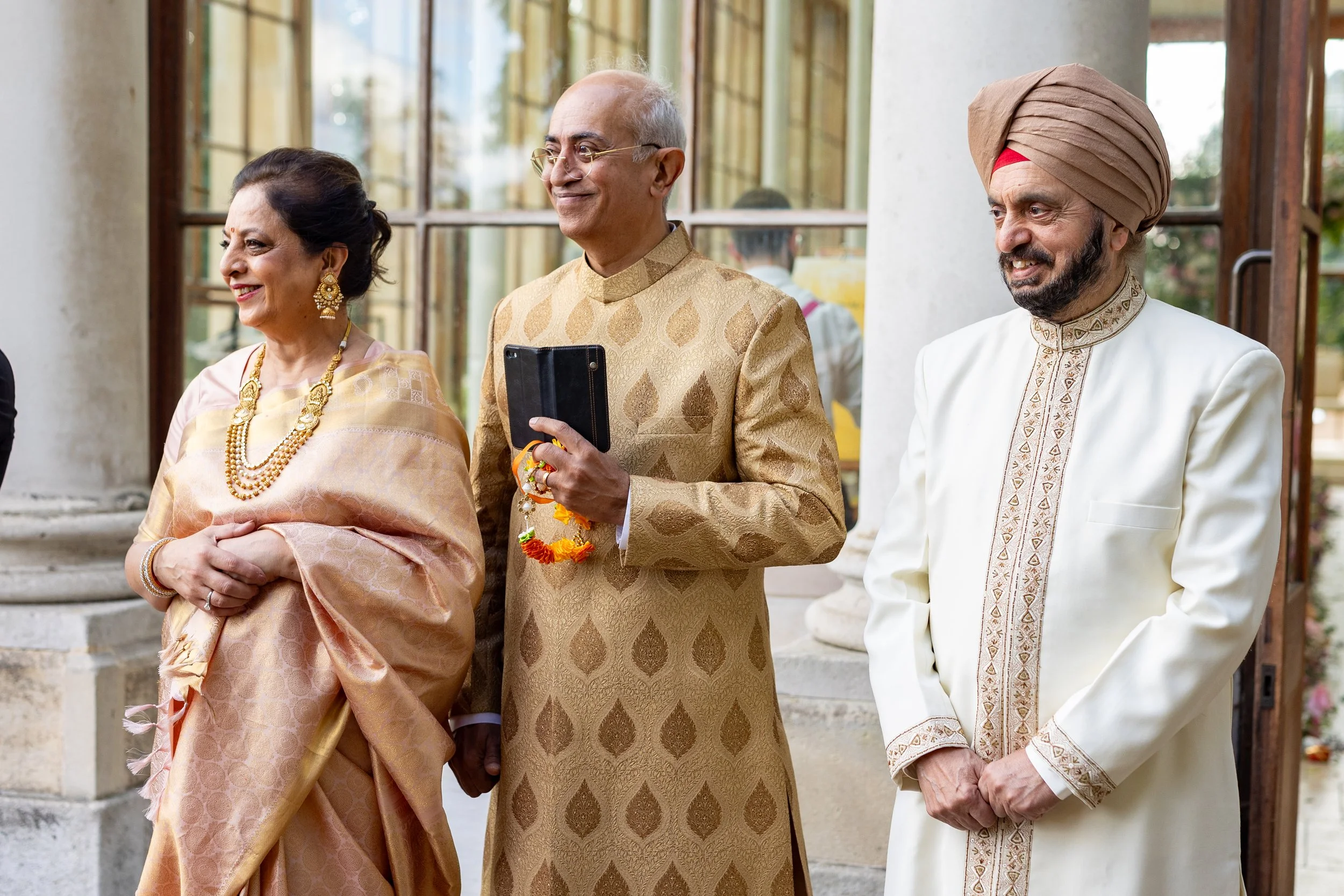 Three distinguished Indian individuals in traditional attire standing outside a building during a celebration or wedding, with one woman and two men smiling.