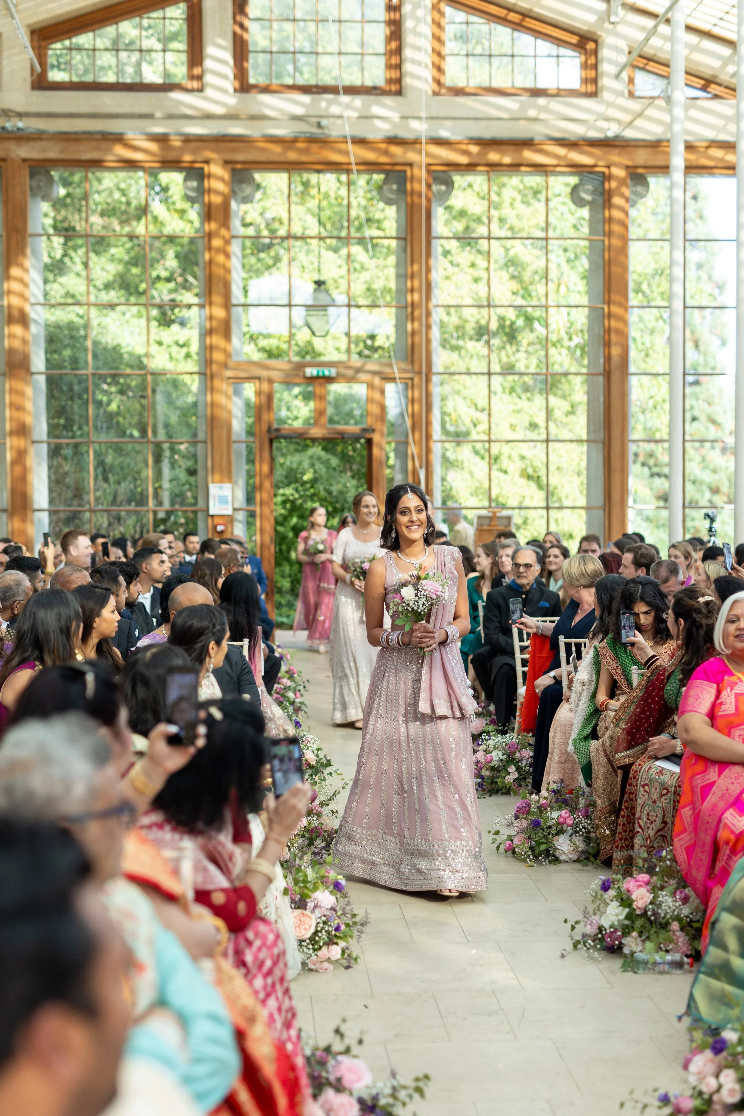A bride in a pink traditional dress walking down the aisle at a wedding ceremony, holding a bouquet and smiling. Guests are seated on either side of the aisle, some are taking photos, in a bright, wooden, glass-walled venue with outdoor greenery visi