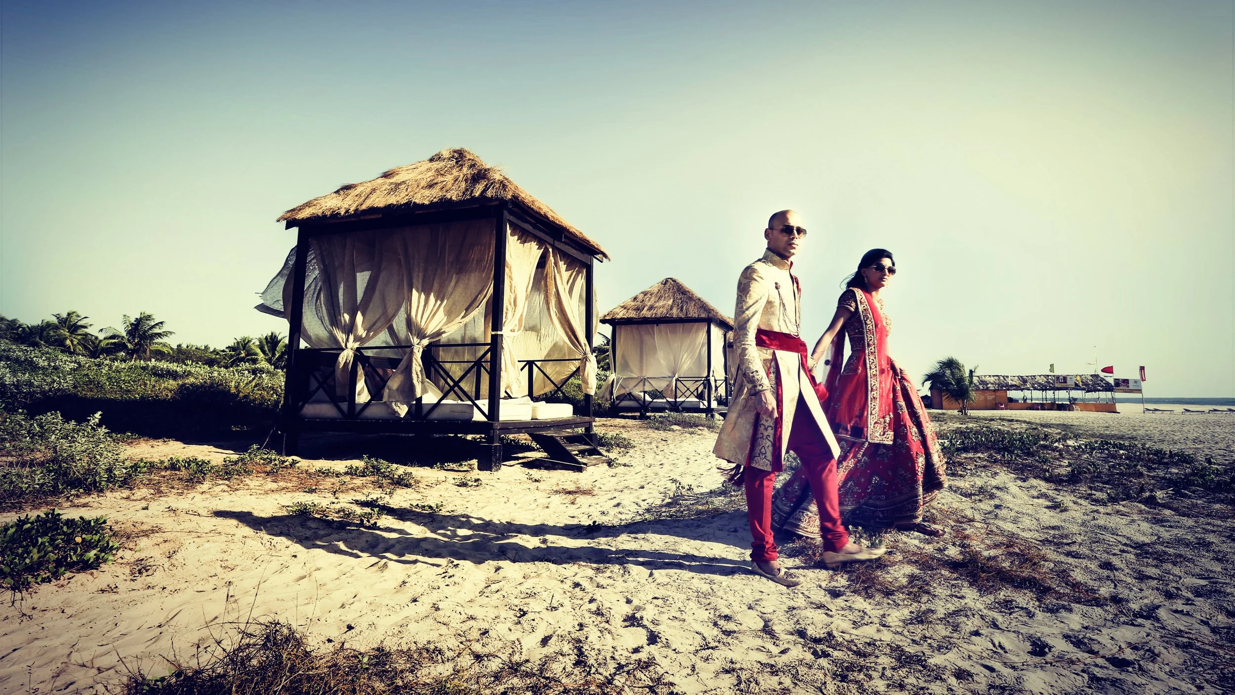 A man and woman in traditional Indian attire walking along a beach with thatched huts and palm trees in the background.