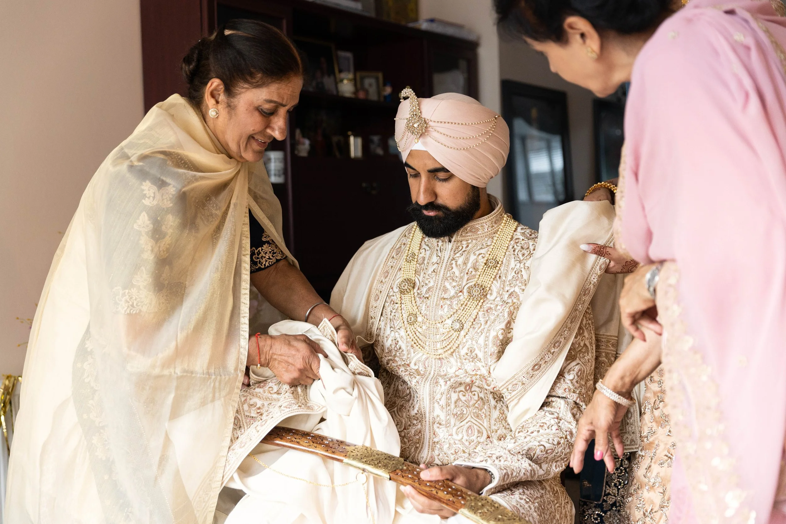 Indian groom in traditional cream and gold sherwani and pink turban, sitting with eyes closed, while family women in pastel sarees assist him during a wedding ceremony.