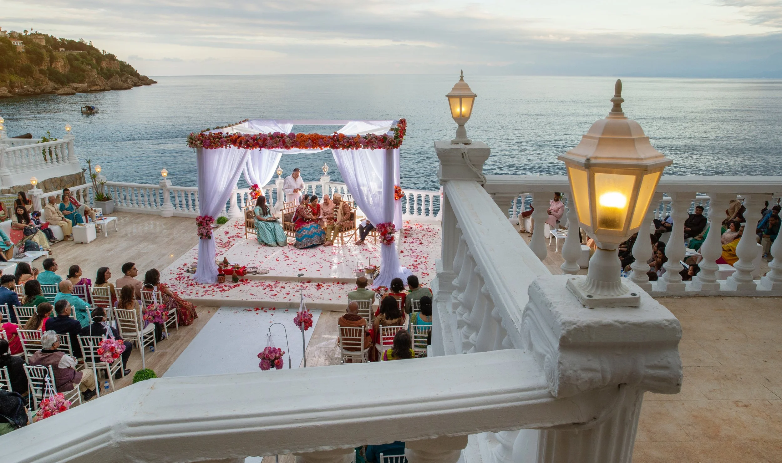 Outdoor wedding ceremony by the sea with a decorated canopy, guests seated on white chairs, and a scenic ocean view at sunset.