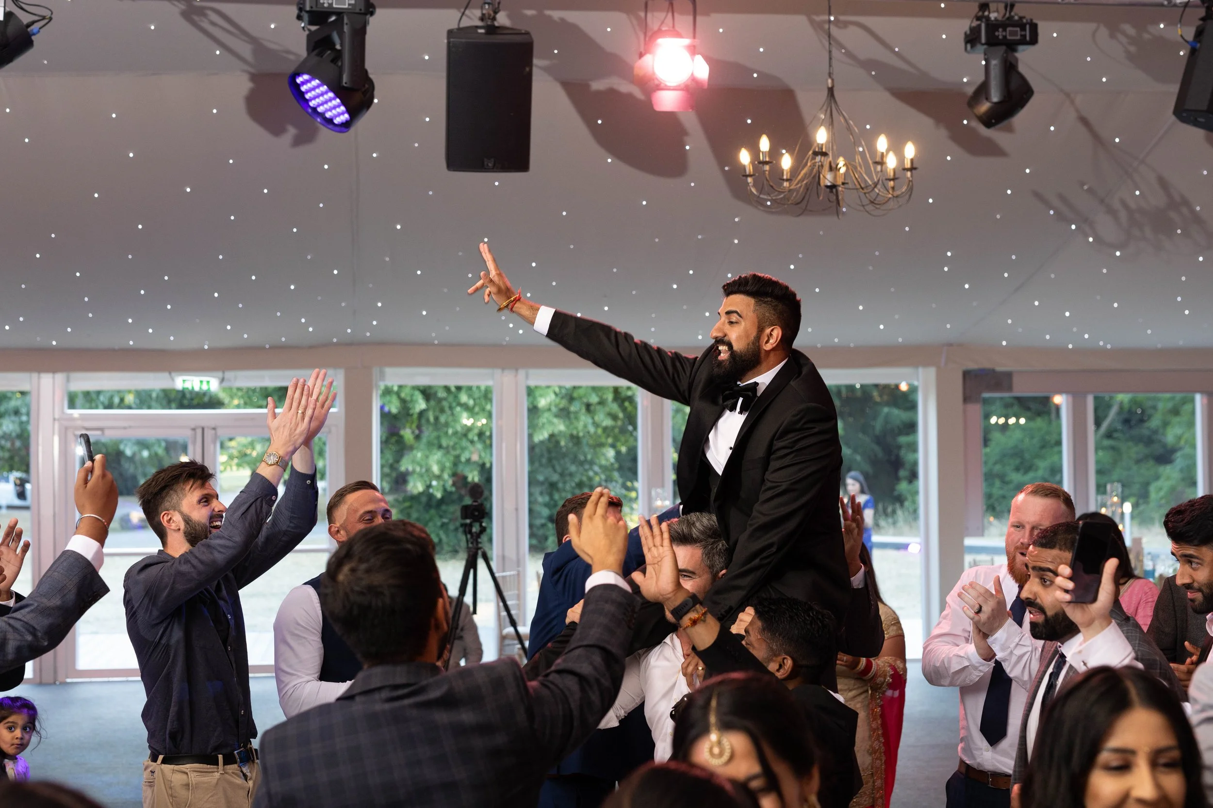 A man in a tuxedo being lifted on people's shoulders during a celebration or wedding reception in a decorated hall with chandeliers and star-like ceiling lights.