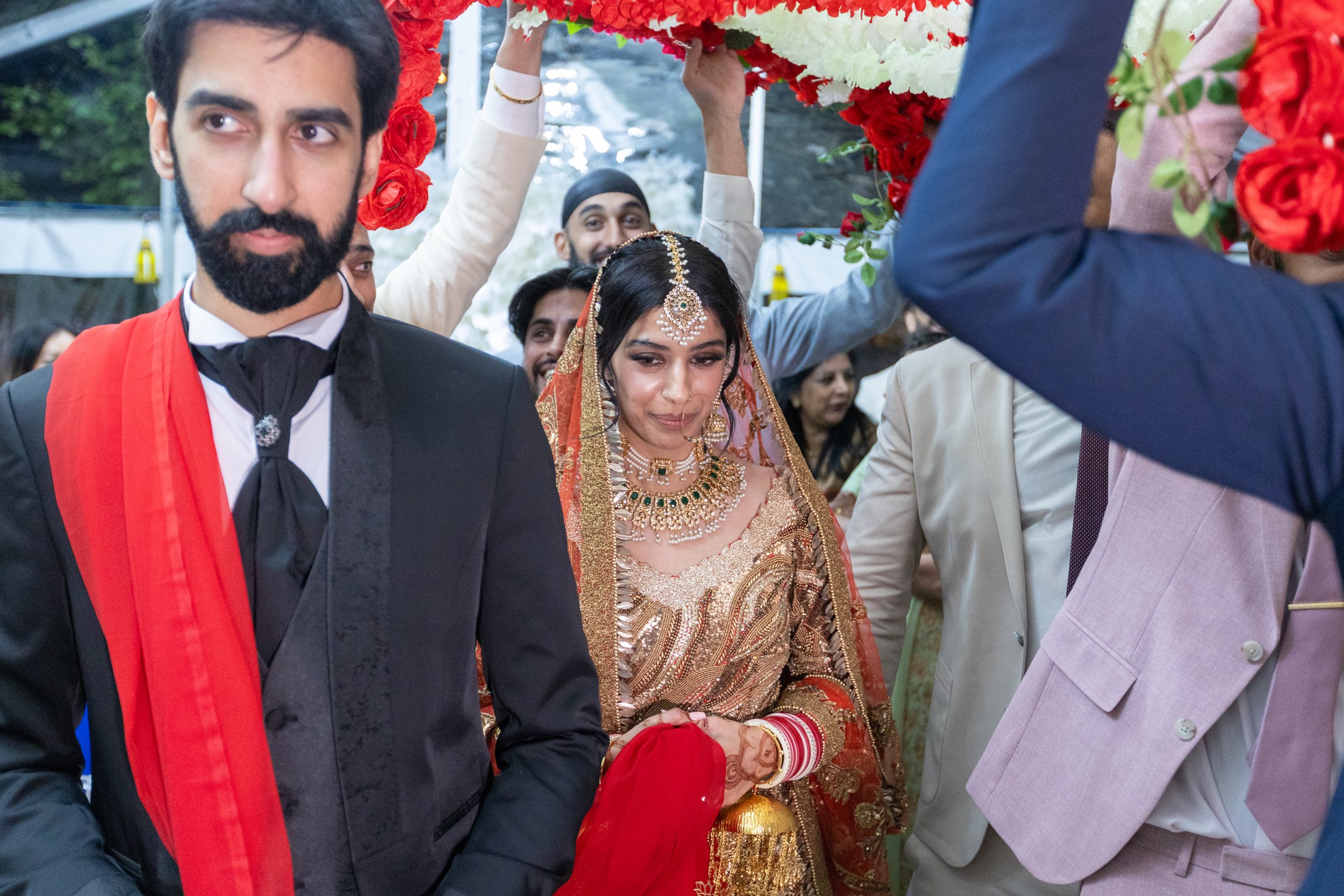 A bride in traditional Indian wedding attire, adorned with jewelry and a red and gold saree, walking under a floral canopy with a groom dressed in a black suit and red stole nearby, surrounded by guests during a wedding celebration.