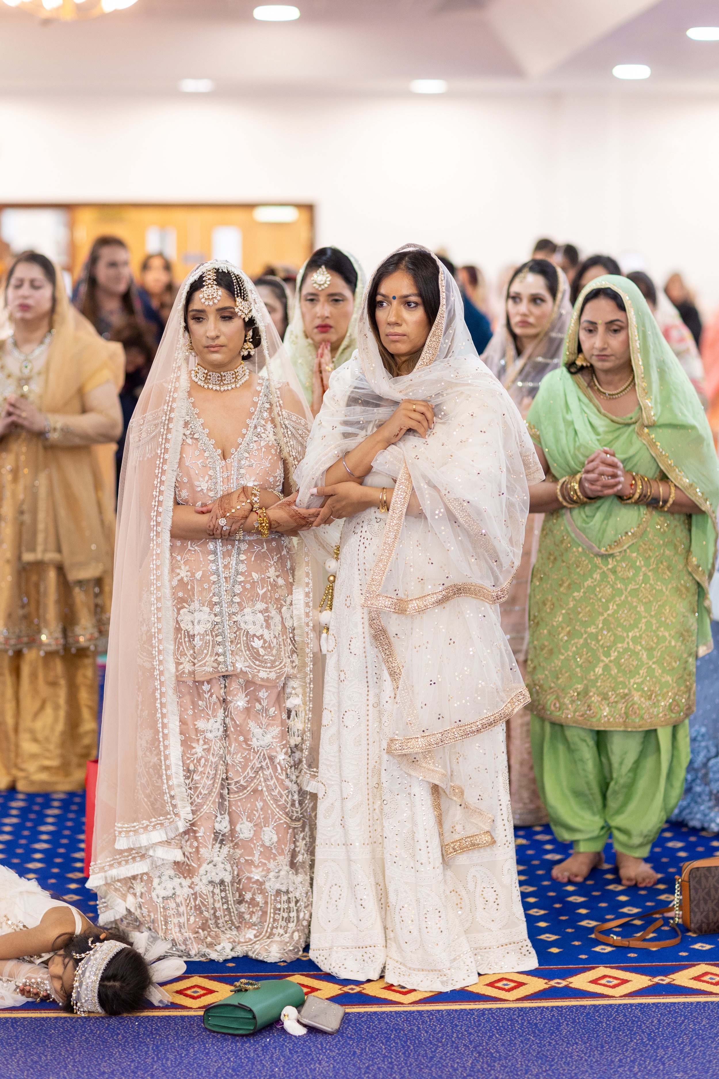 Women participating in a traditional Indian wedding ceremony, dressed in colorful, ornate clothing and jewelry, standing with hands folded in prayer in a decorated indoor venue.