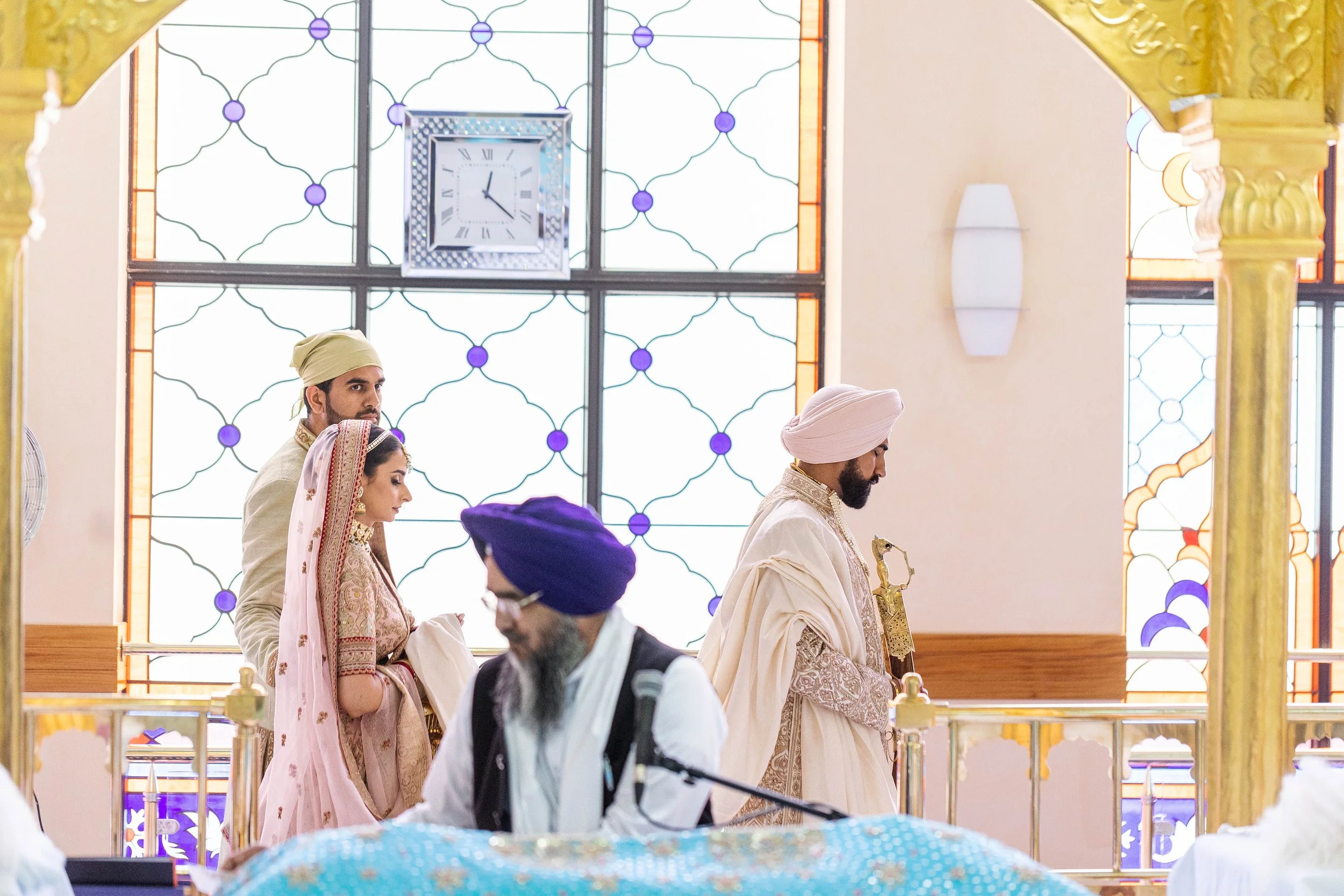Indian wedding ceremony with bride and groom dressed in traditional attire, surrounded by family members in a decorated hall with stained glass windows and a clock.